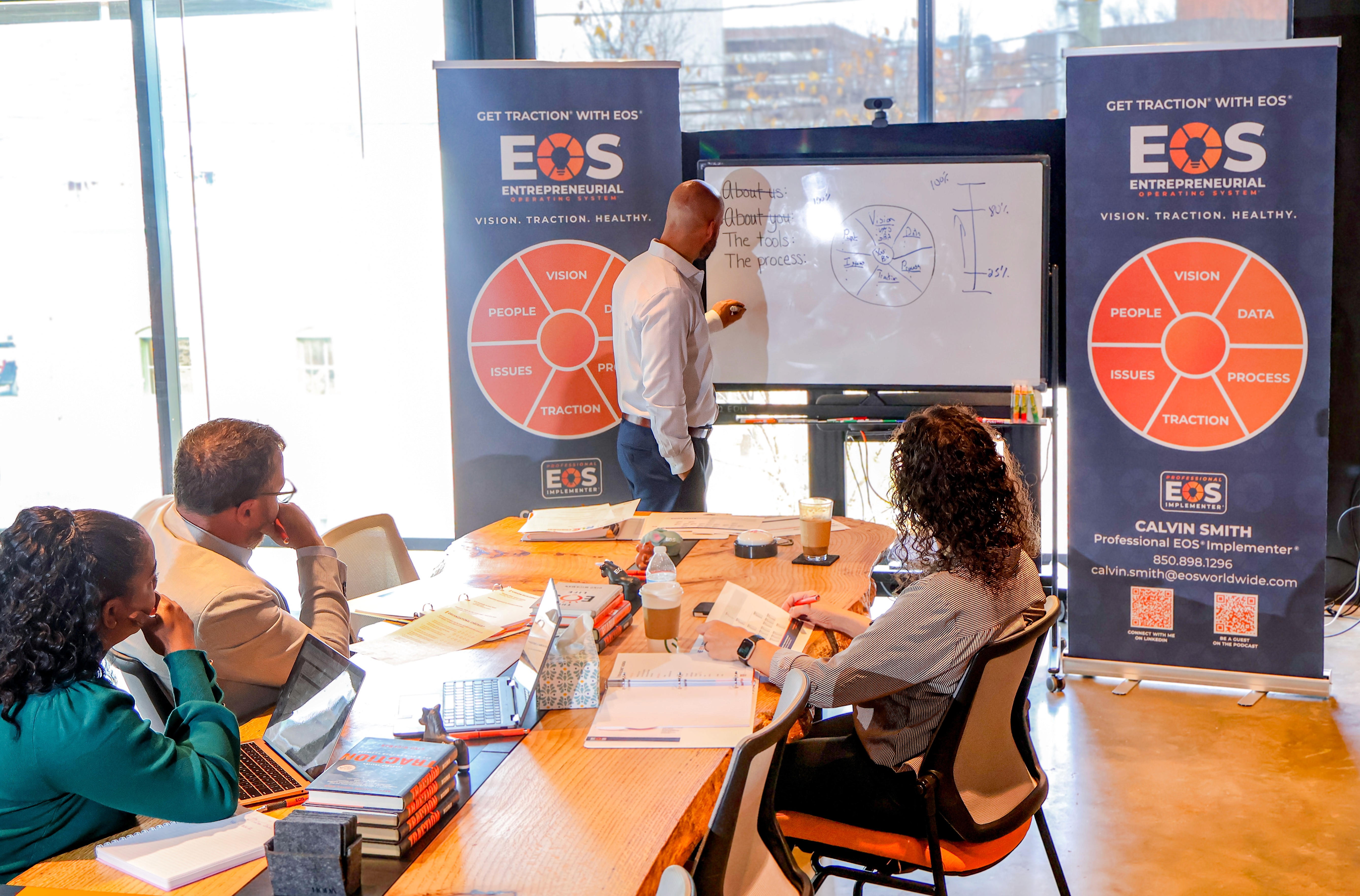 A wooden desk by a window, with sunlight illuminating a laptop, notebook, and a decorative vase.