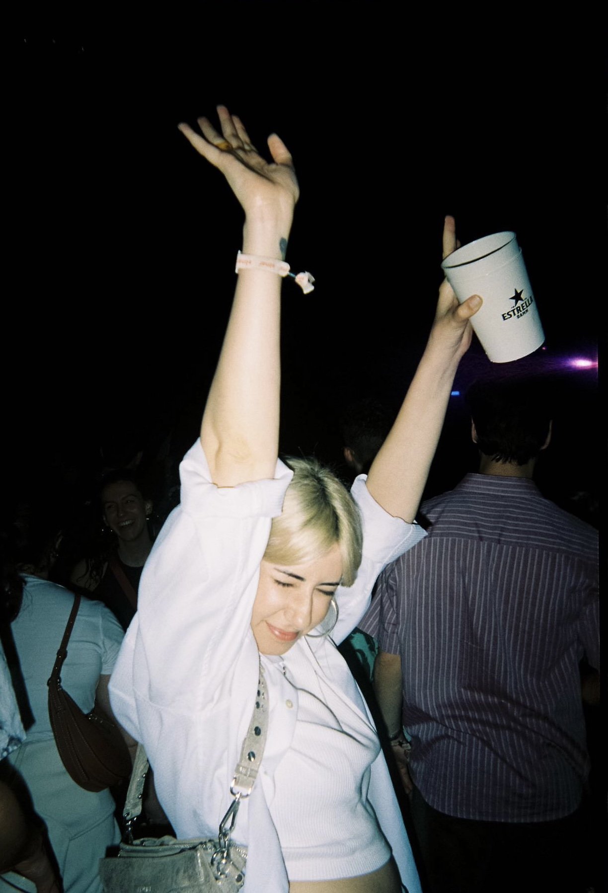 Young woman with short blonde hair dancing or celebrating with eyes closed, holding a white cup, in a crowded dark night setting.