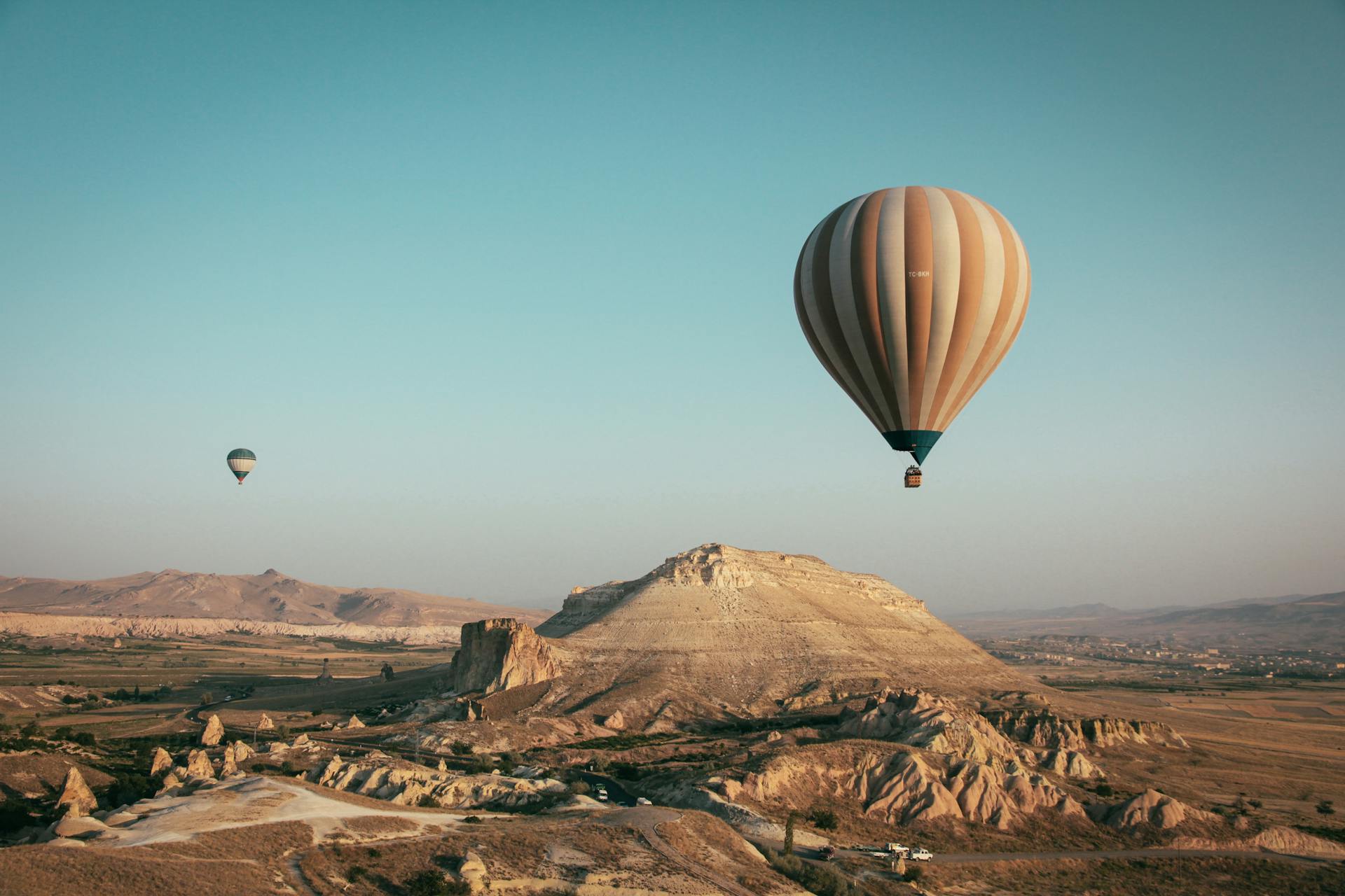 A large, striped hot air balloon flies low over rocky hills and unusual rock formations under a clear blue sky. A second balloon is visible in the distance.