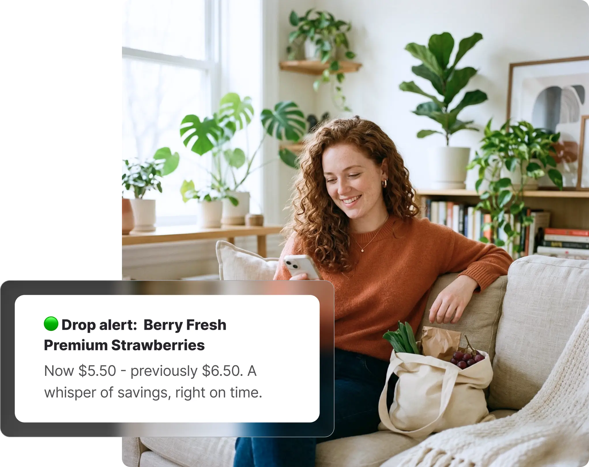 Woman with curly hair sitting on a couch smiling at her phone, with a grocery bag containing fresh produce beside her, receiving a Coles price drop alert for strawberries on Whisprice, with indoor plants in the background.
