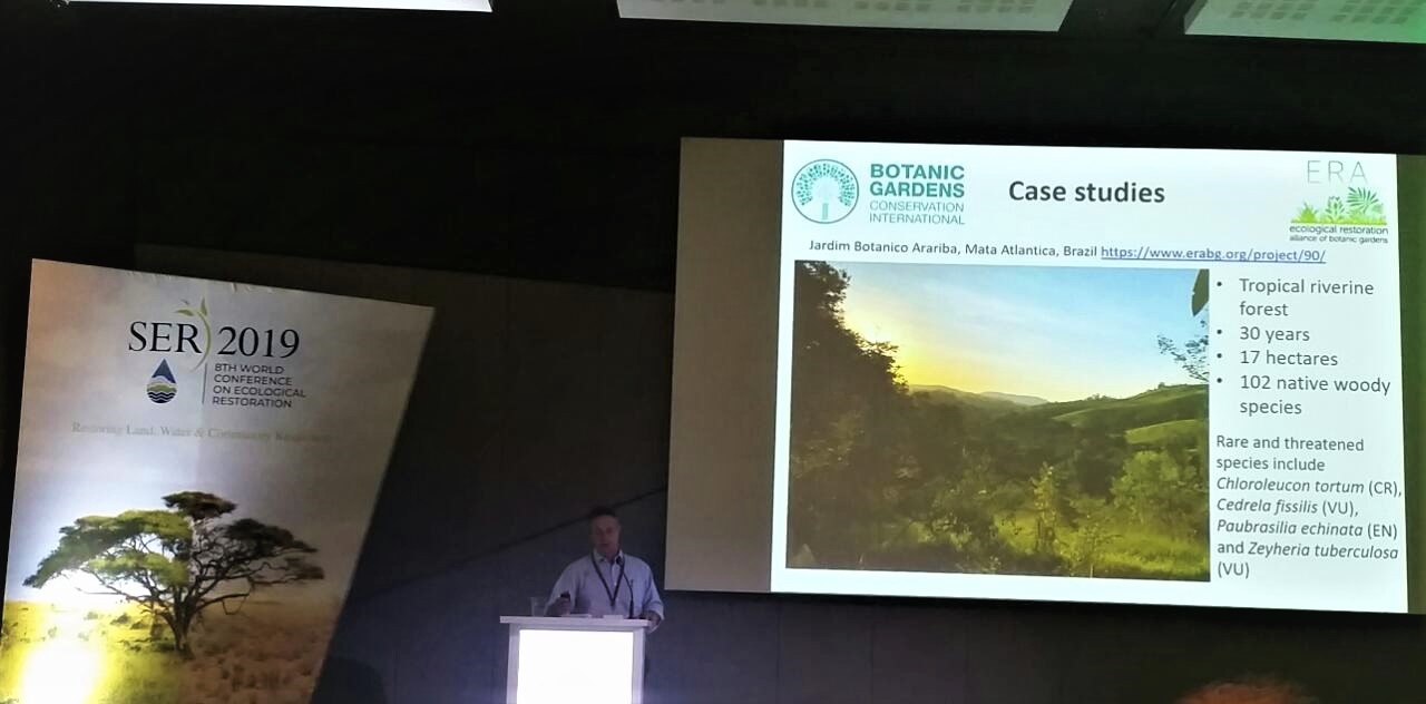 Palestrante no púlpito da 8ª Conferência Mundial de Restauração Ecológica, com slide de estudos de caso ao fundo