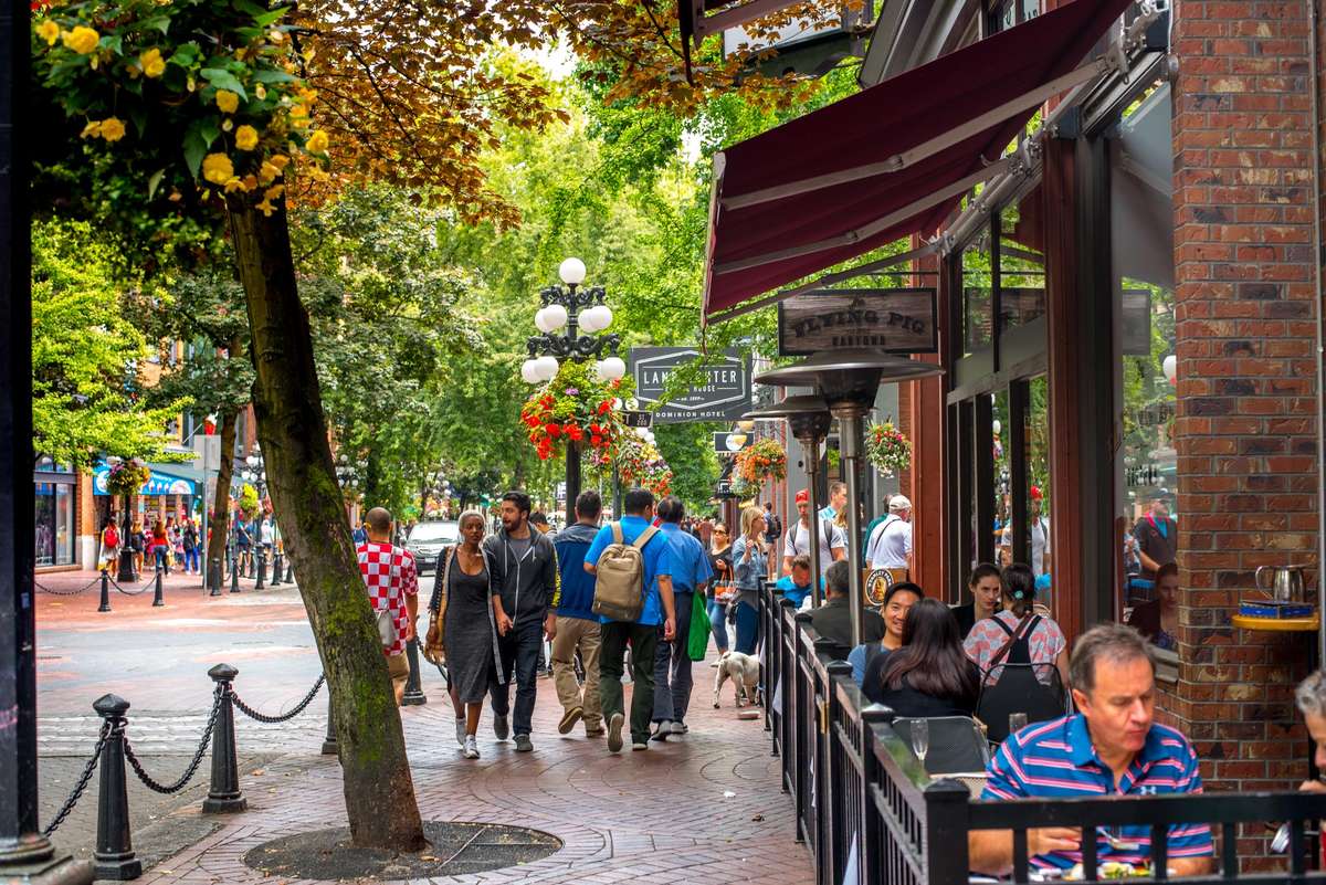 People sit at outdoor cafe tables under a tree with autumn leaves and red awnings on a sunny city sidewalk.