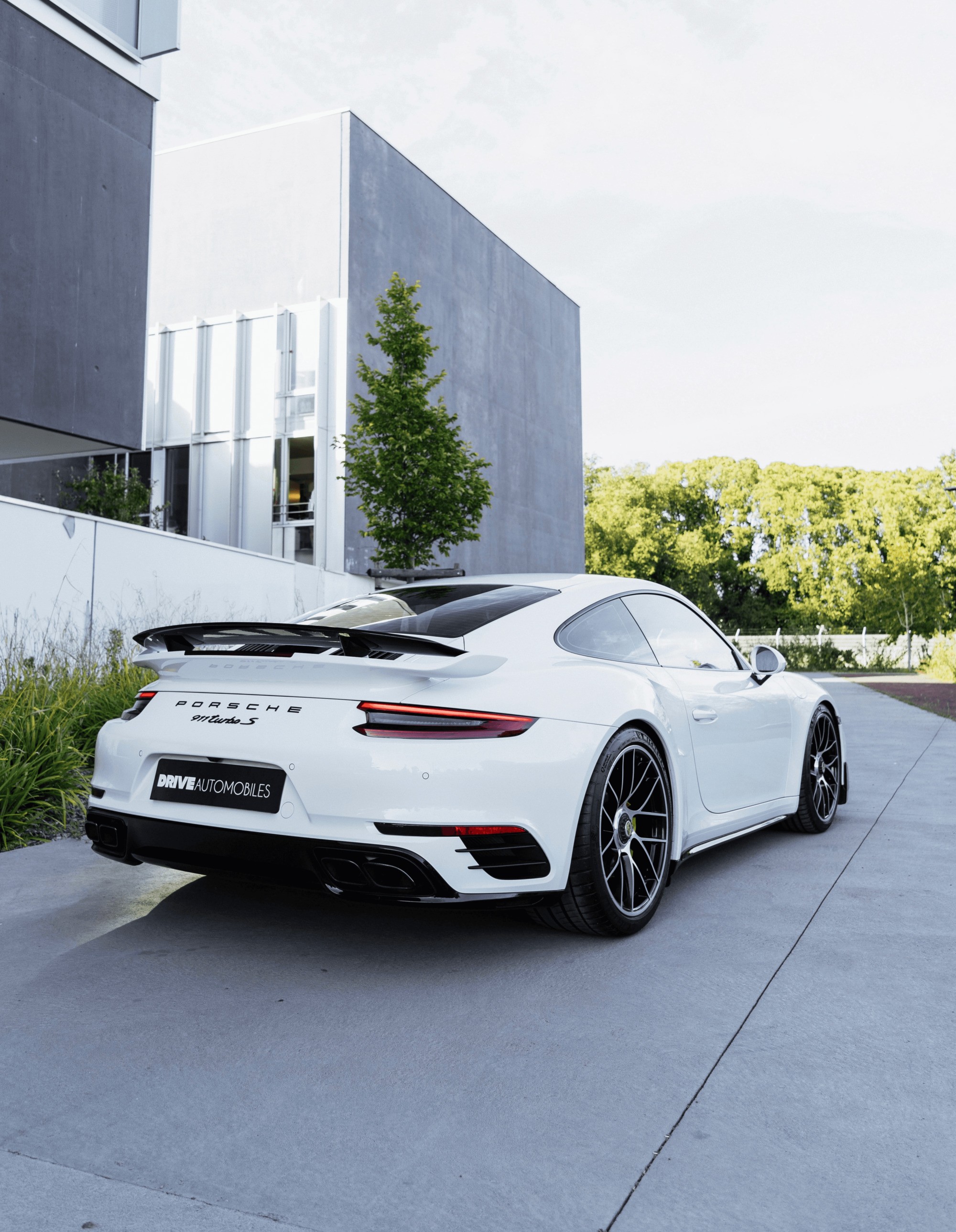 A sleek white sports car, likely a Porsche, is parked on a modern, minimalist driveway with stylish grey pavers, flanked by a contemporary building and lush green trees in the background.