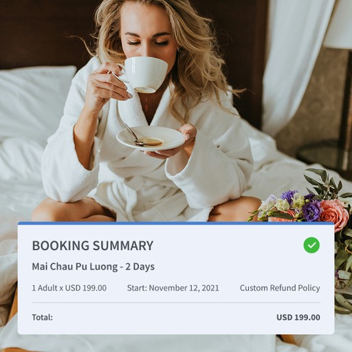 Woman in a white bathrobe sitting on a hotel bed, drinking coffee with a breakfast tray and bouquet of flowers beside her.