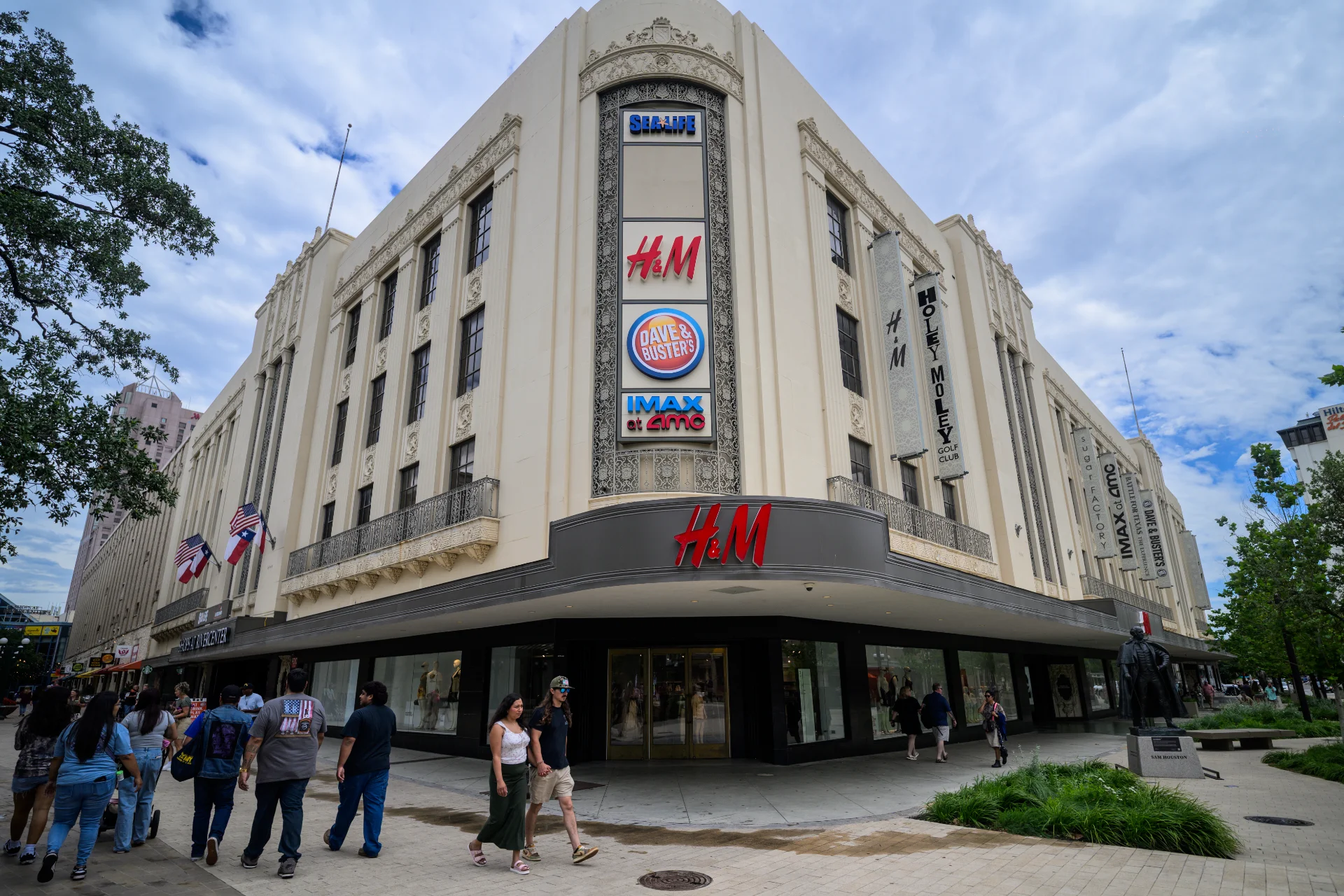 Exterior of the Shops at Rivercenter building with Sea Life, H&M, Dave & Buster's, and IMAX at AMC signage, pedestrians on the sidewalk below