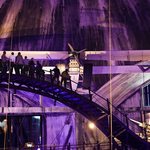 Az emberek egy magasabban fekvő, ívelt hídon sétálnak egy gyengén megvilágított, ipari szerkezetben, amelyet lila fények világítanak meg.