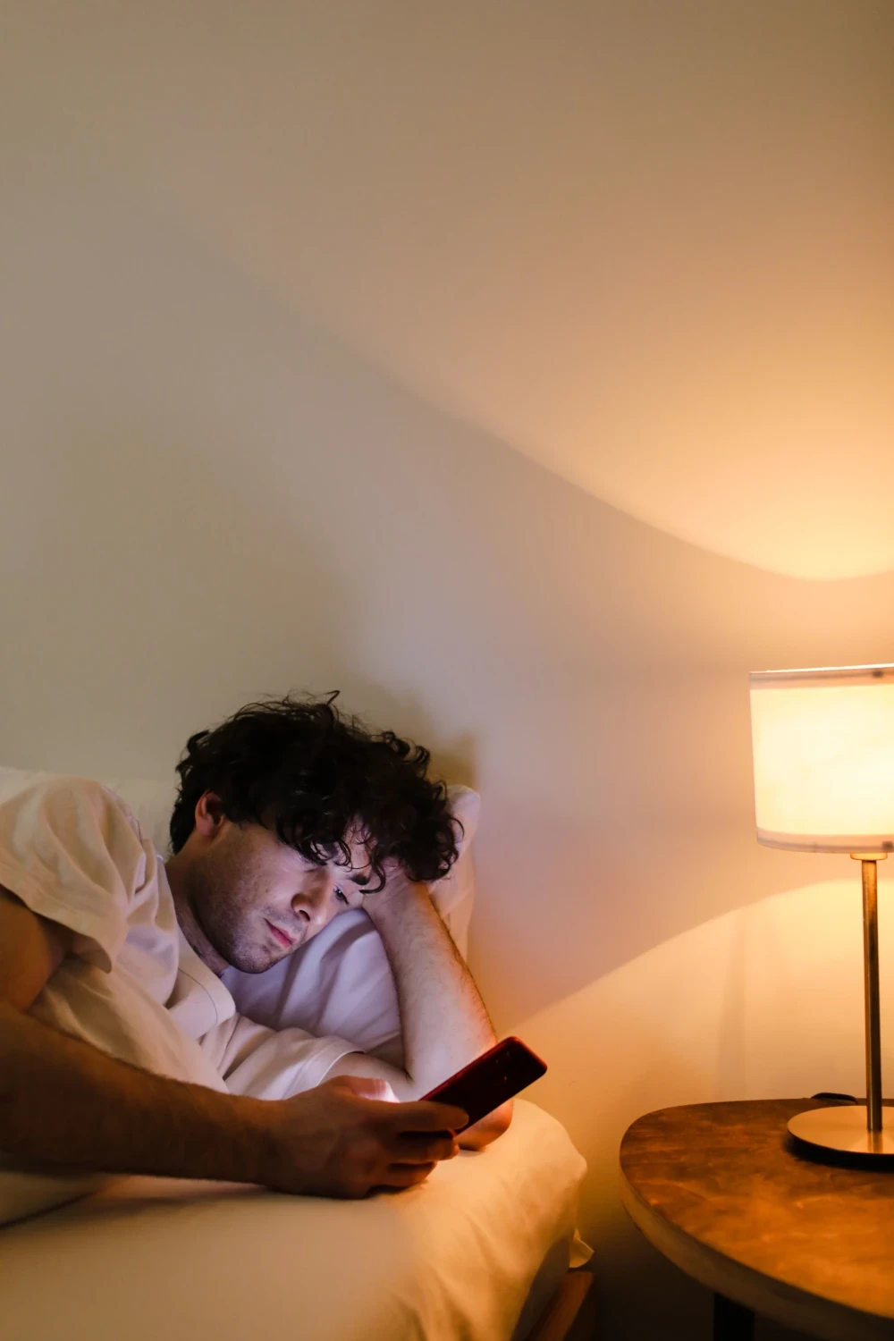 Man lying awake in bed at night, looking at his phone beside a warm bedside lamp.