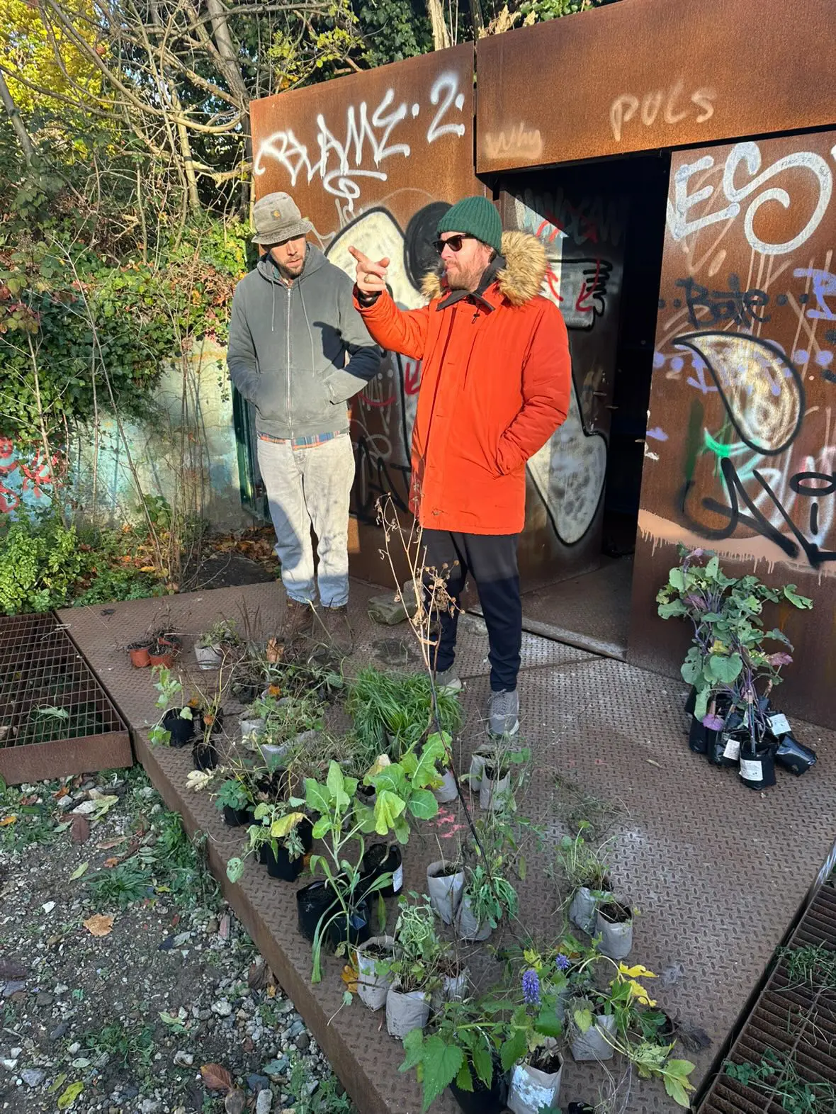 Two people stand in a garden with various plants, smiling and interacting near a wooden structure.