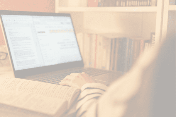 Image of someone on a laptop tying with books i the background and a book on the desk