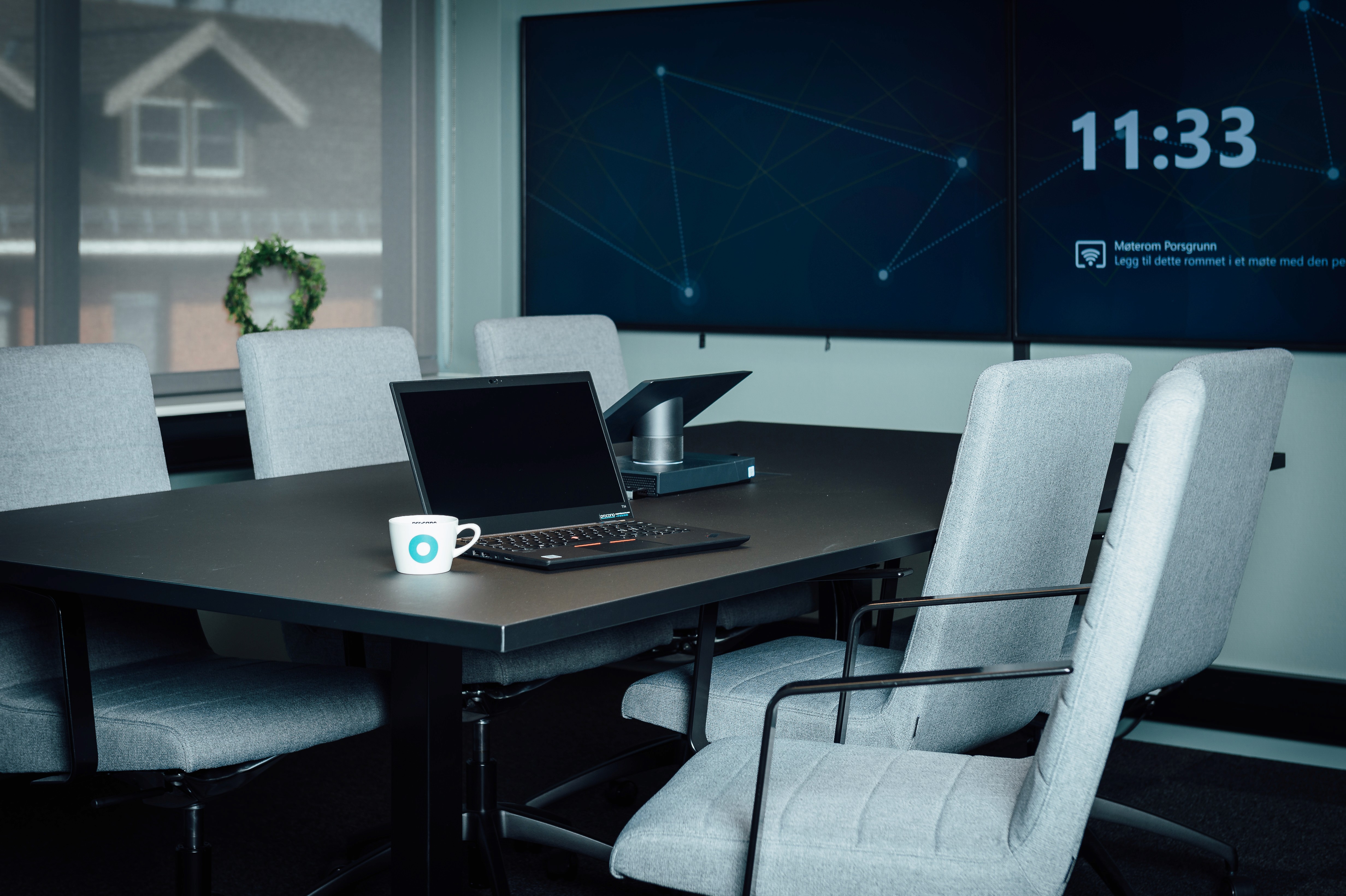 Et moderne møterom med grå møtestoler, et møtebord og en bærbar PC med Procano‑kopp ved siden av. Store skjermer viser et digitalt møteklokkeslett i bakgrunnen.