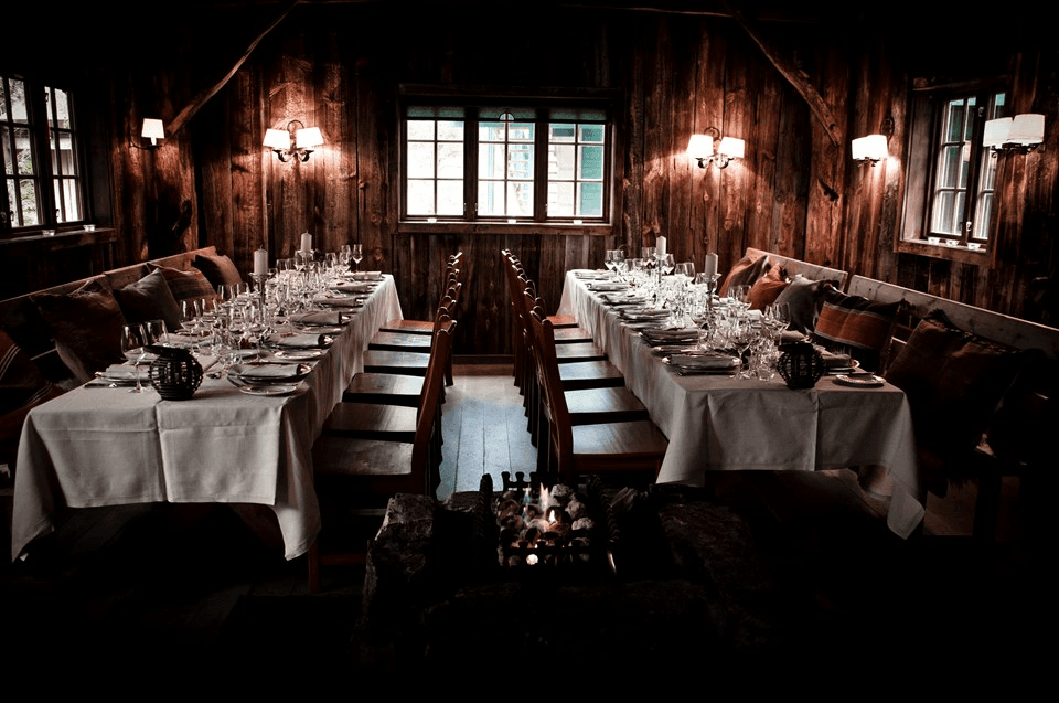 Dimly lit rustic dining room with two long tables covered in white tablecloths, set elegantly with crystal glasses and fine china, surrounded by wooden chairs and soft pillows, creating a warm and inviting atmosphere.