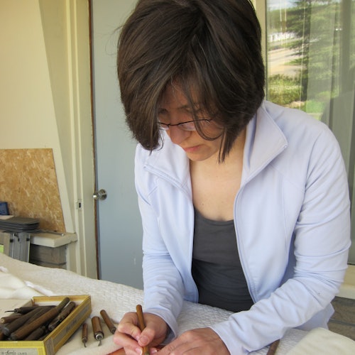 Người có mái tóc ngắn đeo kính và mặc áo khoác màu xanh nhạt, đang tập trung vẽ hoặc viết trên bàn, với nhiều công cụ khác gần đó.