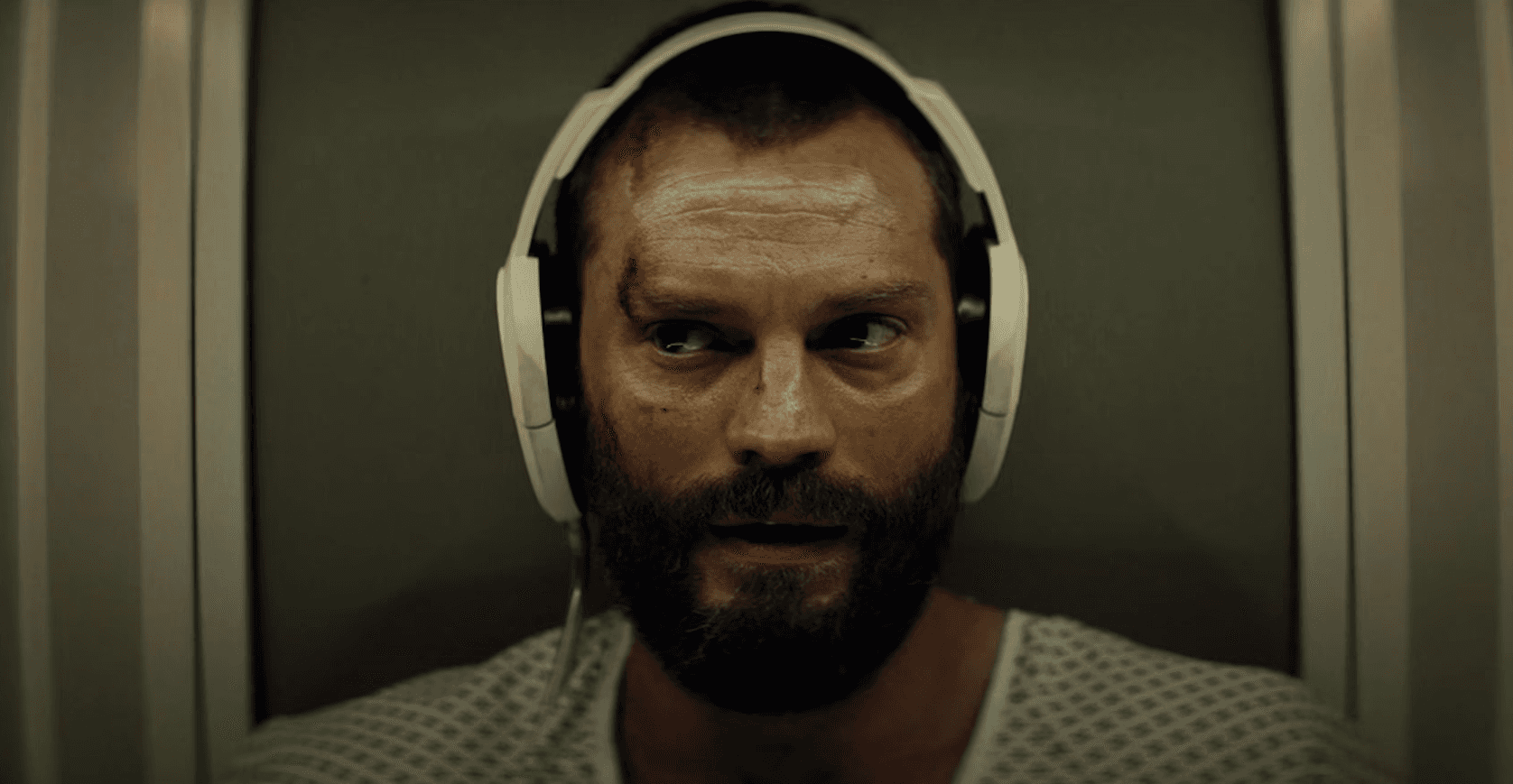 A man with a beard and headphones stares intently, appearing contemplative in a dimly lit space.