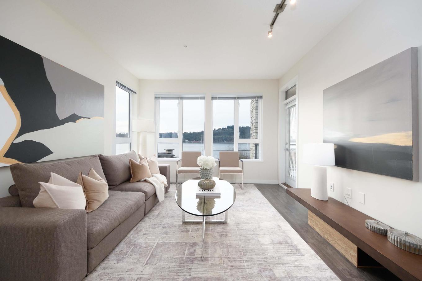 A modern living room featuring a sofa, coffee table, artwork, and large windows with natural light.