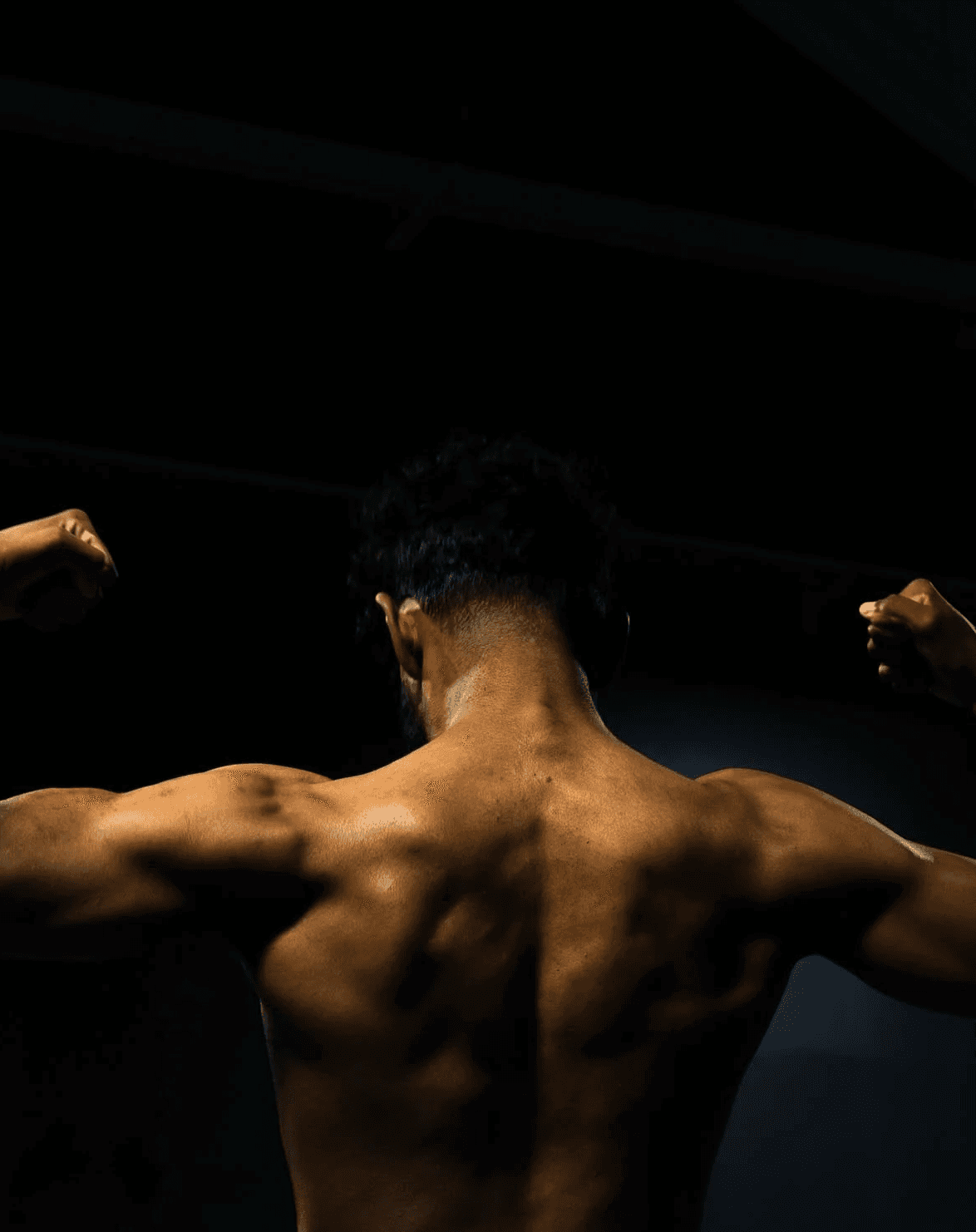 Back view of a male athlete flexing his muscles under low lighting in a gym.
