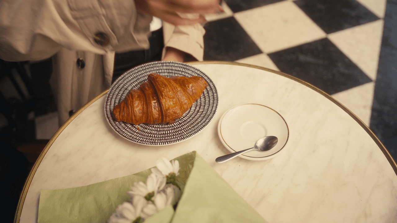 A croissant on a table set for breakfast, next to a cup of coffee and a glass of orange juice, with a checkered floor in the background