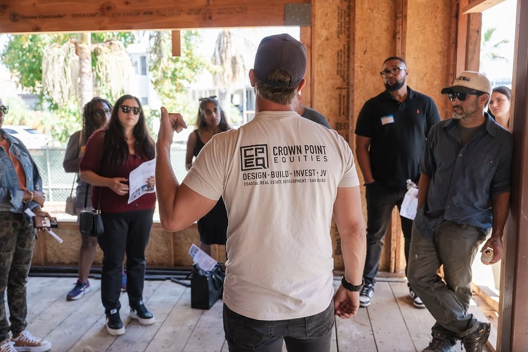 A close-up of a small, unusual house with a geodesic-like dome shape. Its roof is covered in a thick layer of green grass and moss, blending into the grassy hills behind it. The walls are a dark material, and the front features a large, triangular window design with white frames. The sky is hazy and overcast.