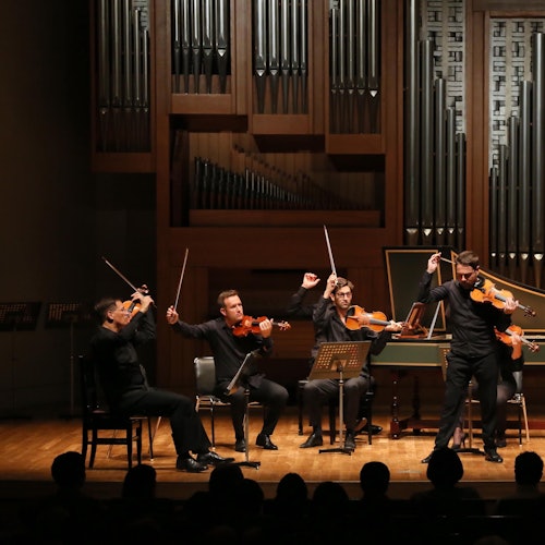 Four musicians performing on stage with string instruments in front of an organ; audience silhouettes visible in the foreground.