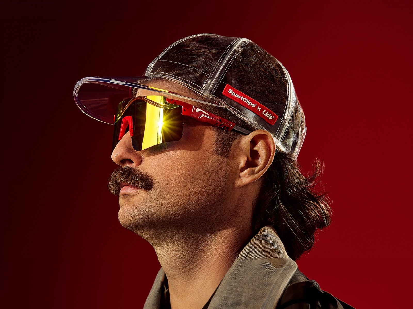 Person wearing a backward cap and yellow-tinted visor sunglasses photographed against a vibrant red background, creating a bold promotional portrait with dramatic lighting.