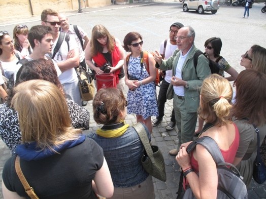 A man giving a lecture to a group of people in an ancient stoned square.