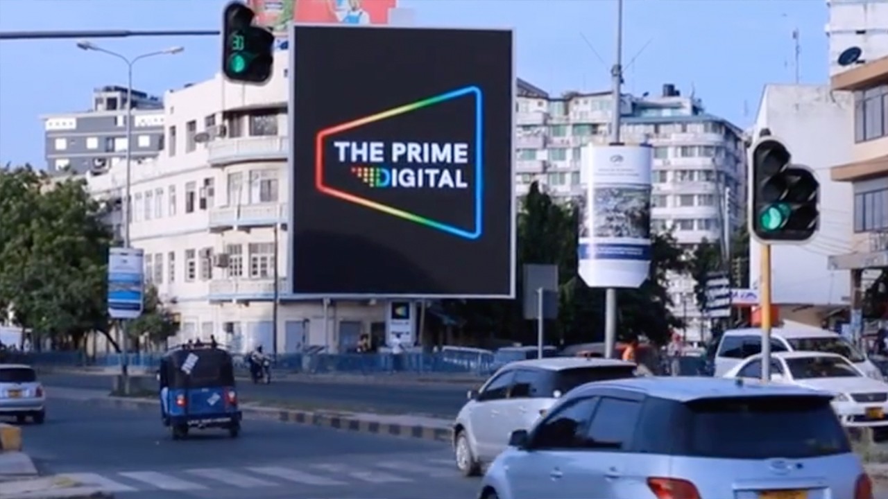 Sunlight streaming through lush green trees, casting shadows on a modern architectural building with glass panels and vertical wooden slats.
