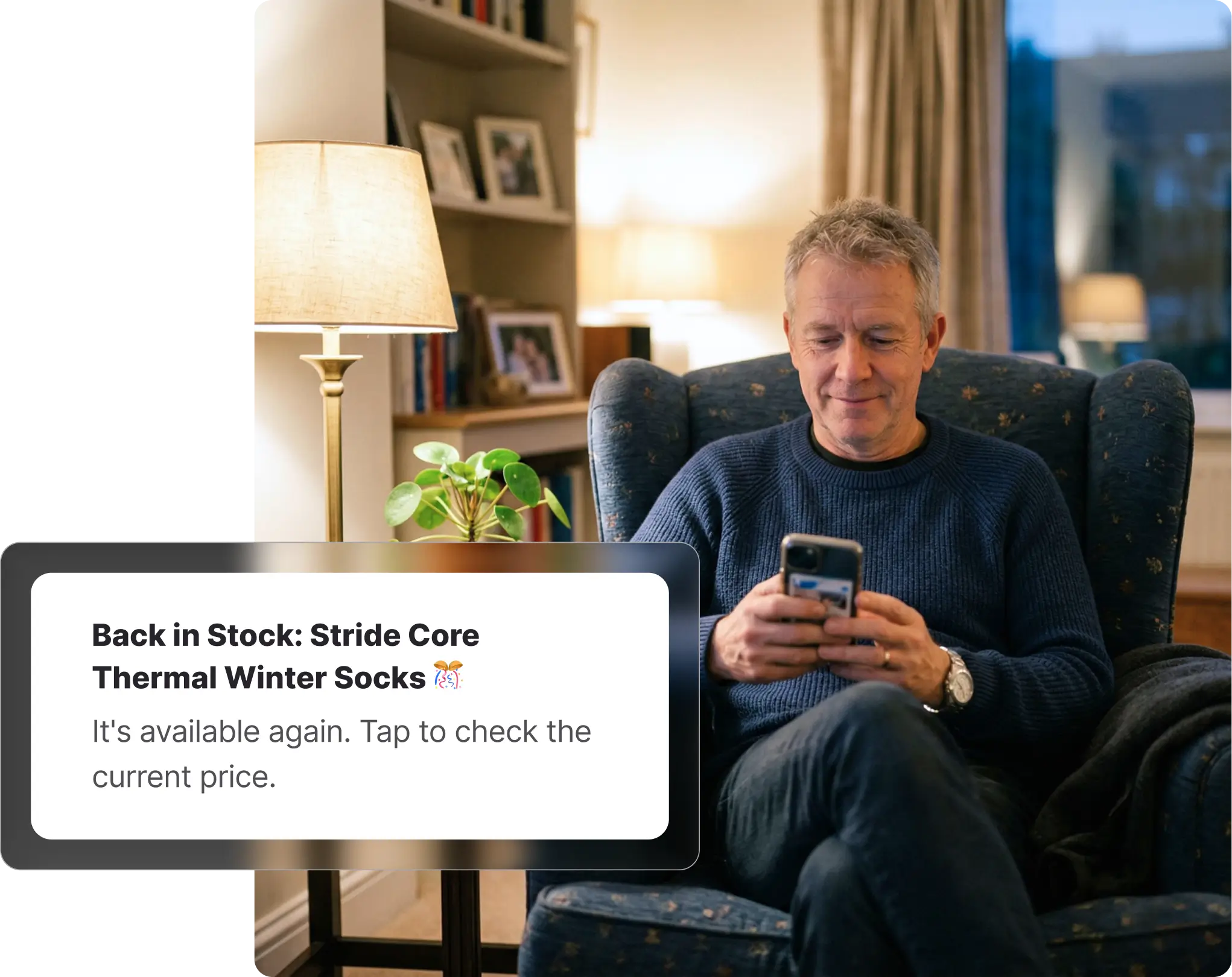 Older man sitting in an armchair at home checking his phone, receiving a BIG W back-in-stock notification, with a bookshelf and lamp in the background.