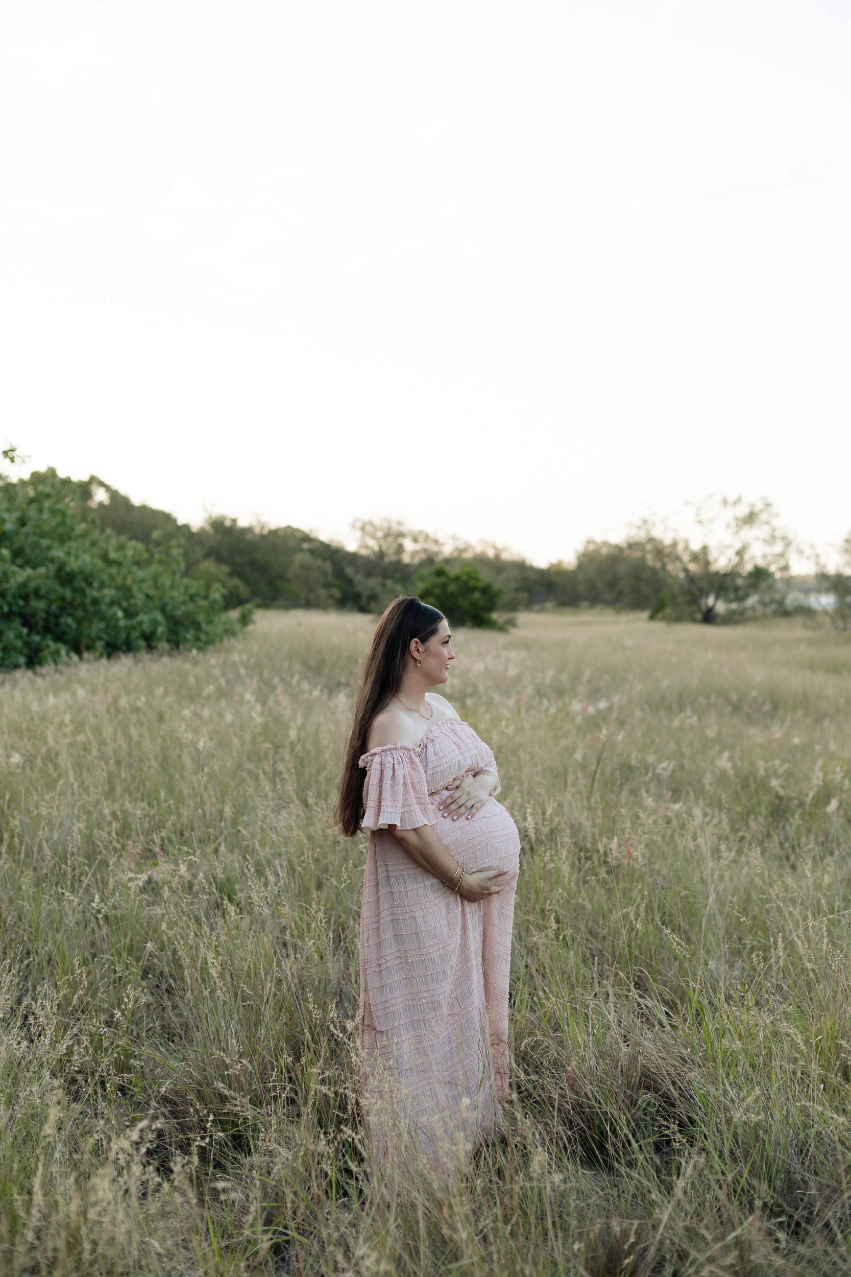Editorial maternity photo of pregnant woman in flowing dress surrounded by tall grass at sunset in North Queensland