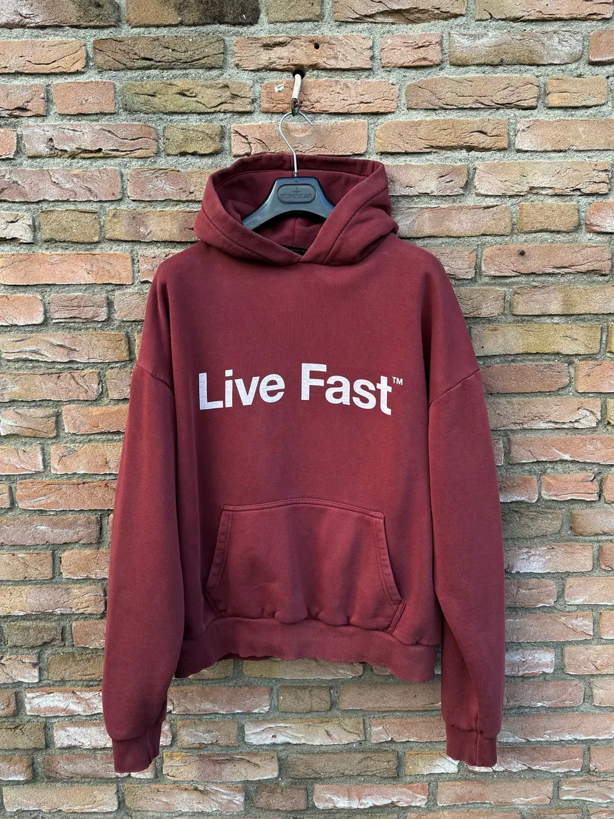 Bordeauxroter Live Fast Hoodie mit weißem Frontprint, hängend vor einer Backsteinwand – Unisex Streetwear Pullover für Fashion-, Lifestyle- und E-Commerce-Produktdarstellungen.