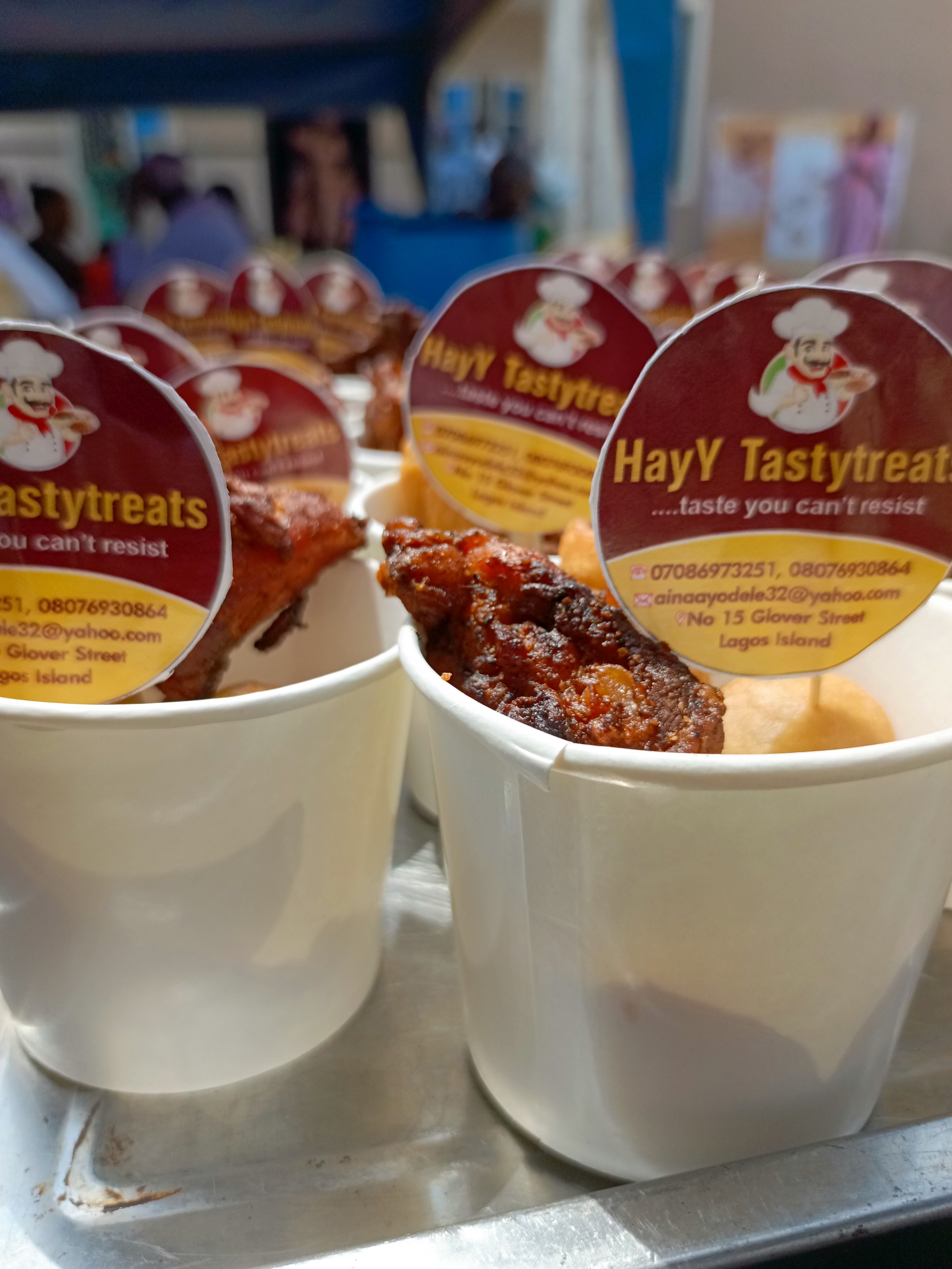 Close-up of several small white cups containing fried chicken pieces and a creamy sauce. Each cup has a round HayY Tastytreats label.
