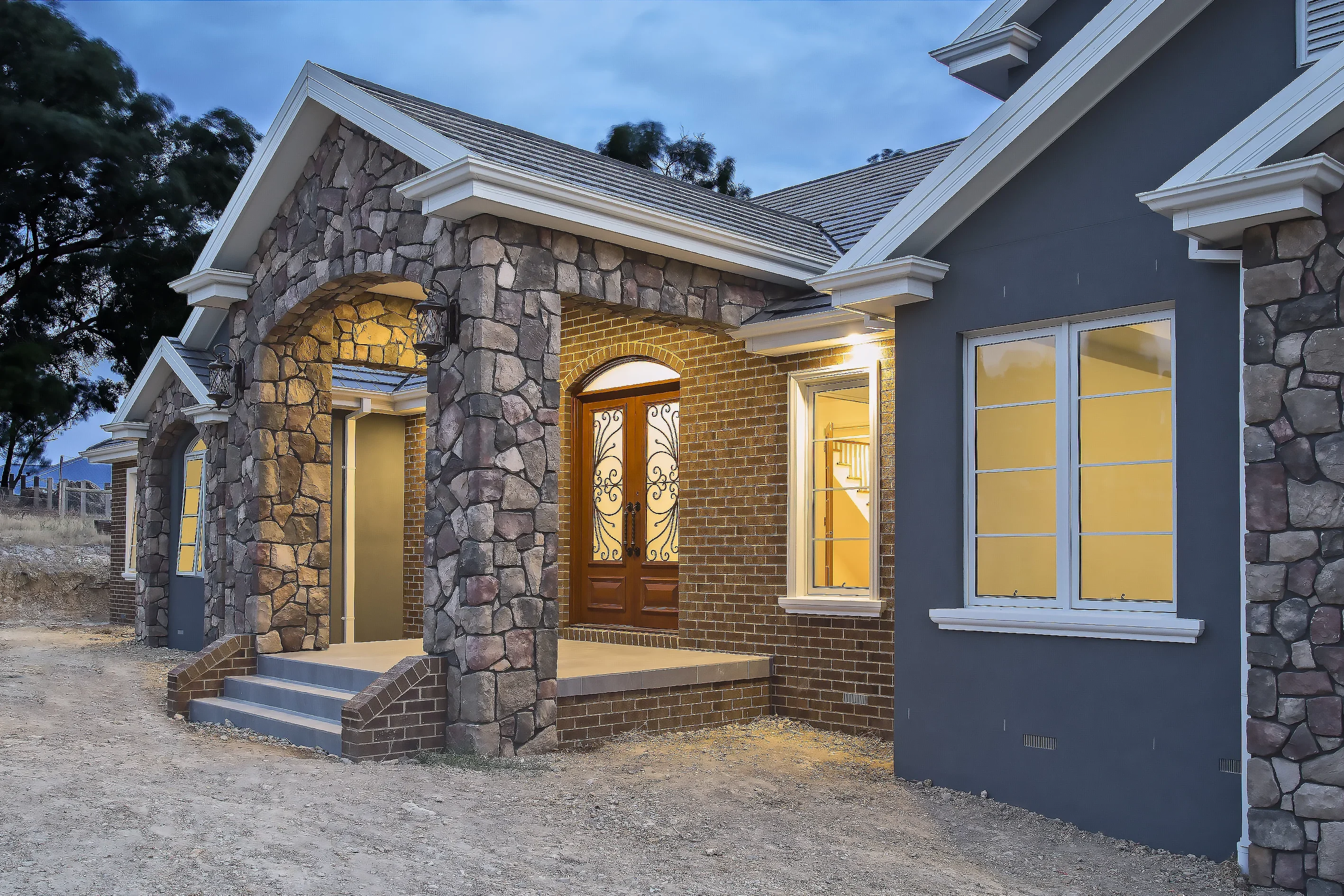 Stone archway entry porch of Plenty Residence with timber double doors, brick walls and illuminated windows at dusk