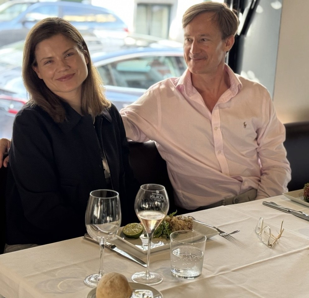 Tosca de Jong sitting at a restaurant table with her partner, smiling warmly. She wears a dark navy jacket; he wears a pale pink shirt.