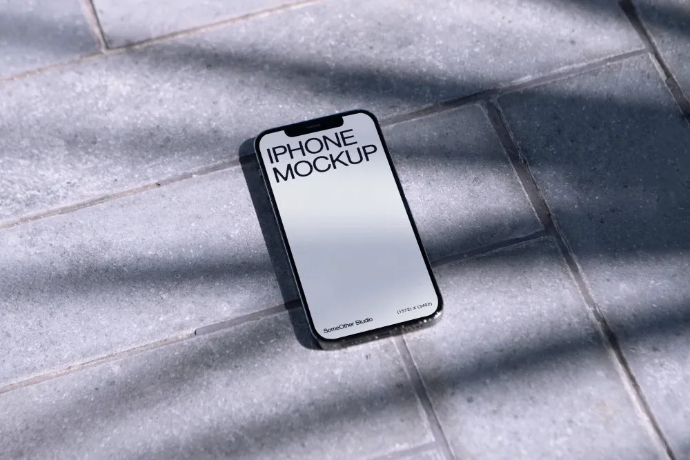 An iPhone mockup placed on gray outdoor tiles, with soft shadows from nearby foliage creating a relaxed, natural ambiance in bright sunlight.