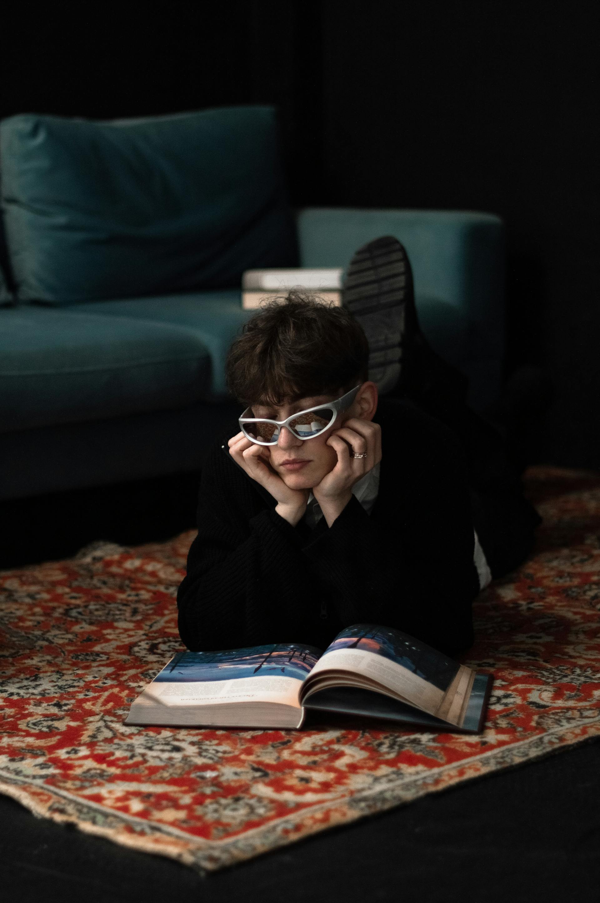 A young person wearing silver sports sunglasses lies on their stomach on a patterned rug, resting their chin on their hands while looking at an open magazine.