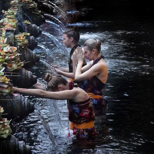 三位身著色彩繽紛沙龍裙的女性正在流動的水泉下站立並祈禱，水泉上裝飾著供品。