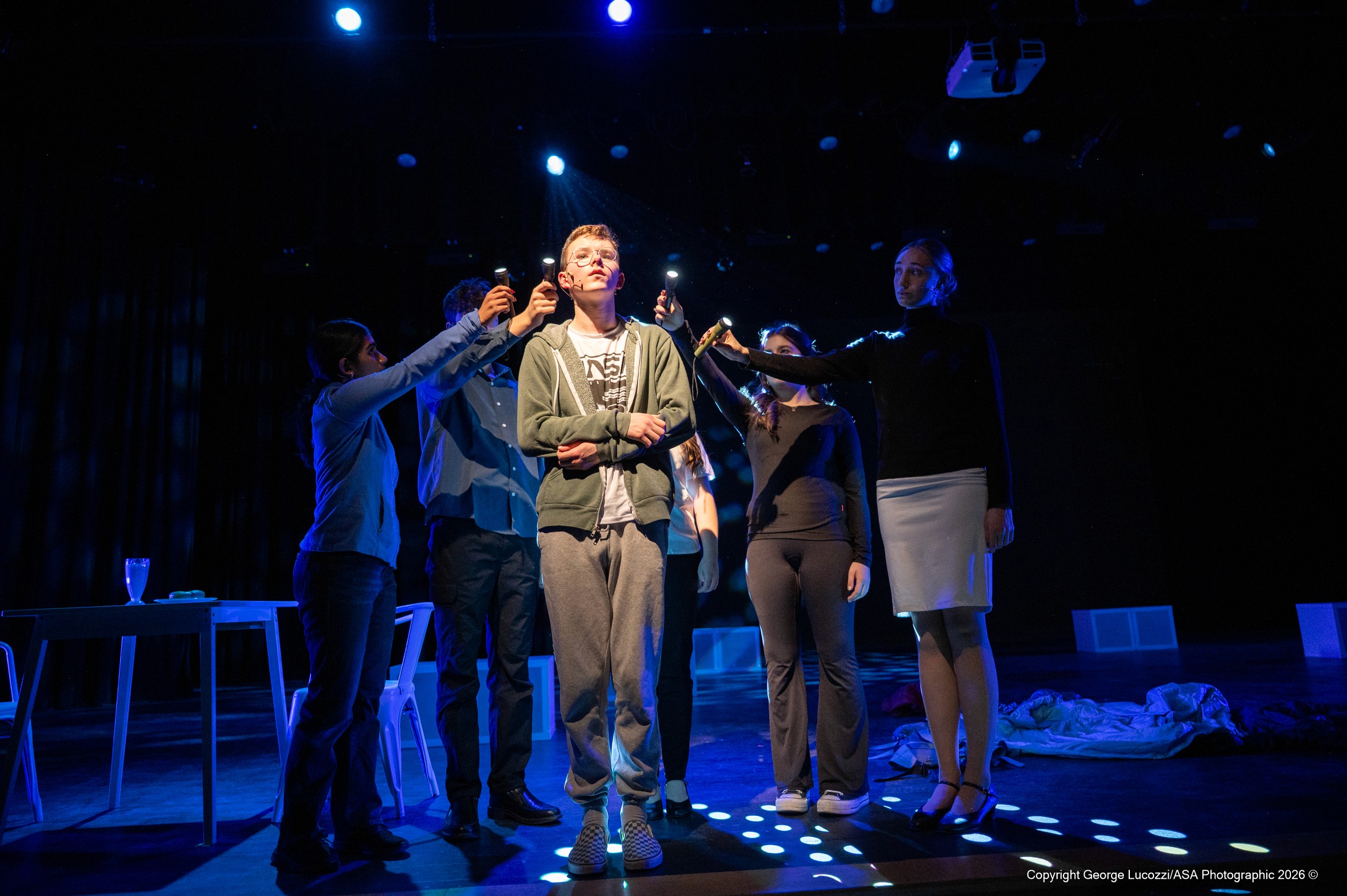 At the stage edge, Christopher gazes outward in a clean followspot while the “Voices” surround him, aiming flashlights into the distance; blue top light and cool white dotted gobo effects heighten the sense of scale and anticipation.