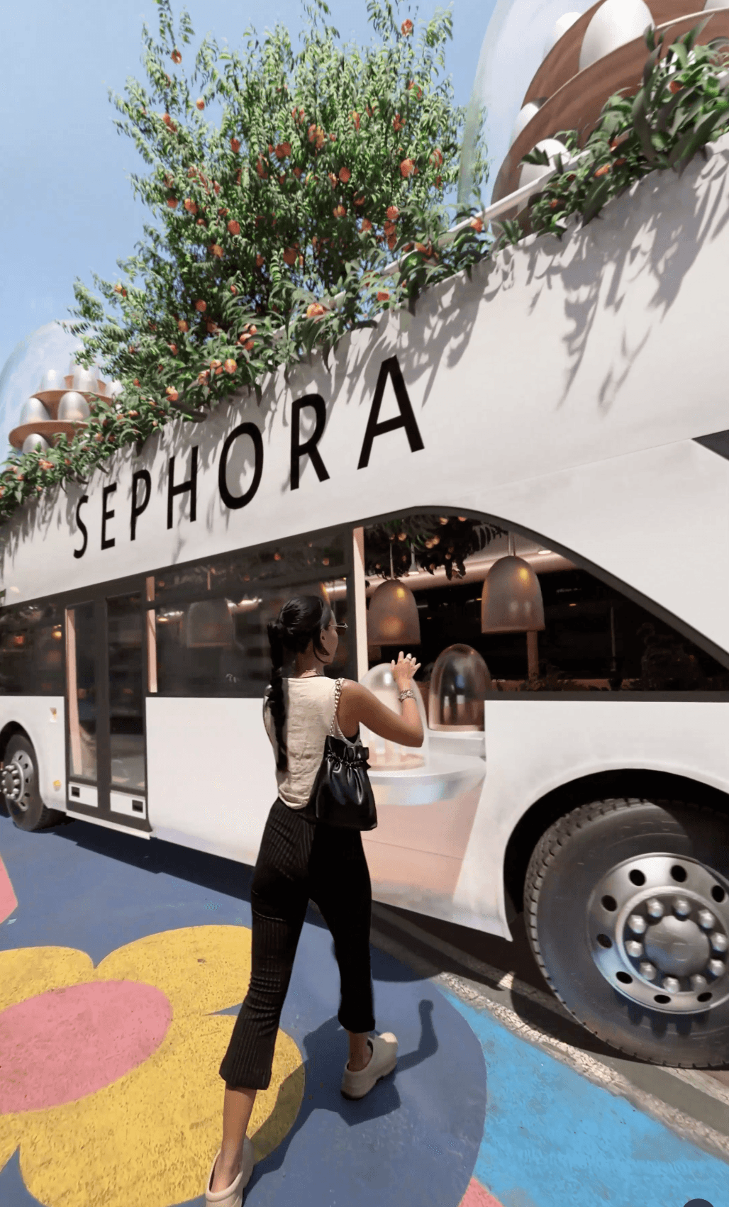 A woman interacts with a Sephora-branded bus overflowing with lush greenery and orange fruit