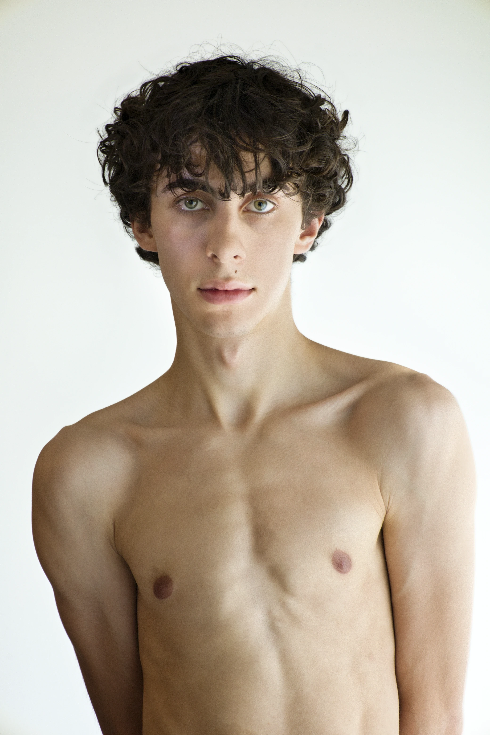 A young person with short, curly hair, standing shirtless against a light background, looking directly at the camera.