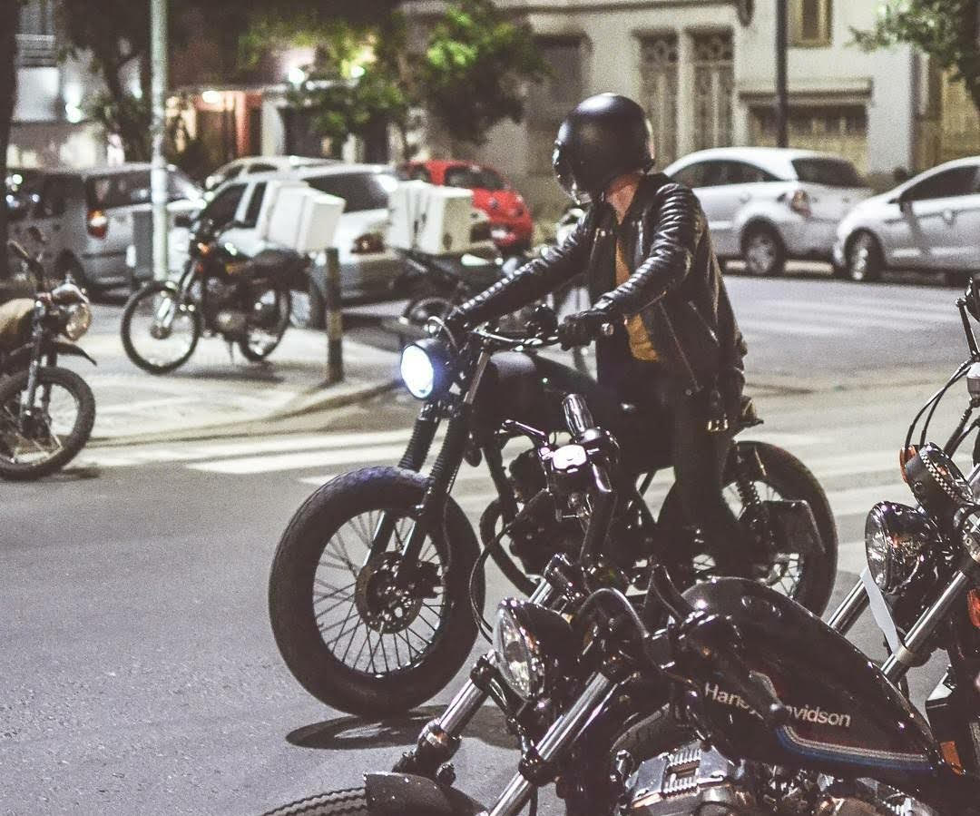 David wearing a black helmet and leather jacket rides a motorcycle at night in an urban setting. Several other motorcycles and parked cars are visible in the background.