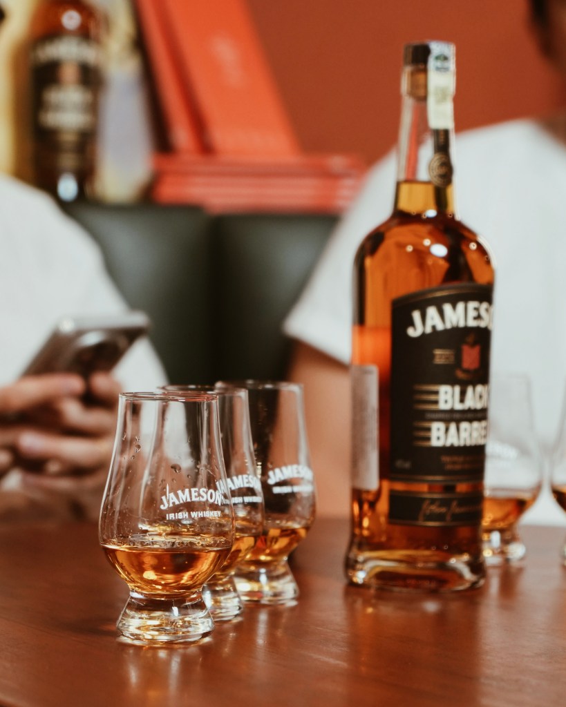 A bottle of Jameson Black Barrel whiskey with three glasses filled with whiskey on a wooden table, alongside a blurred person holding a smartphone.