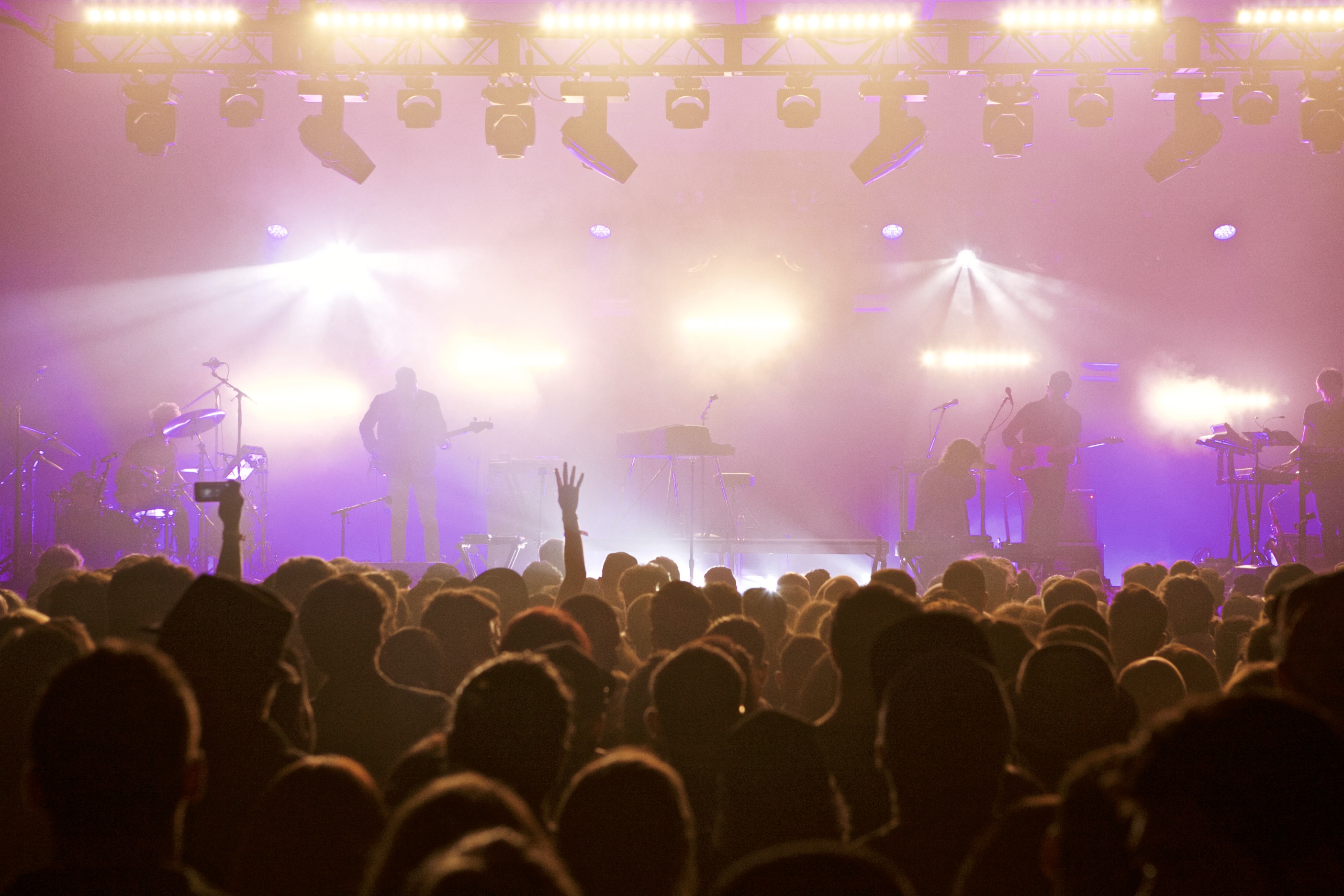Wide crowd view of a concert with the band onstage and bright lights washing the room.