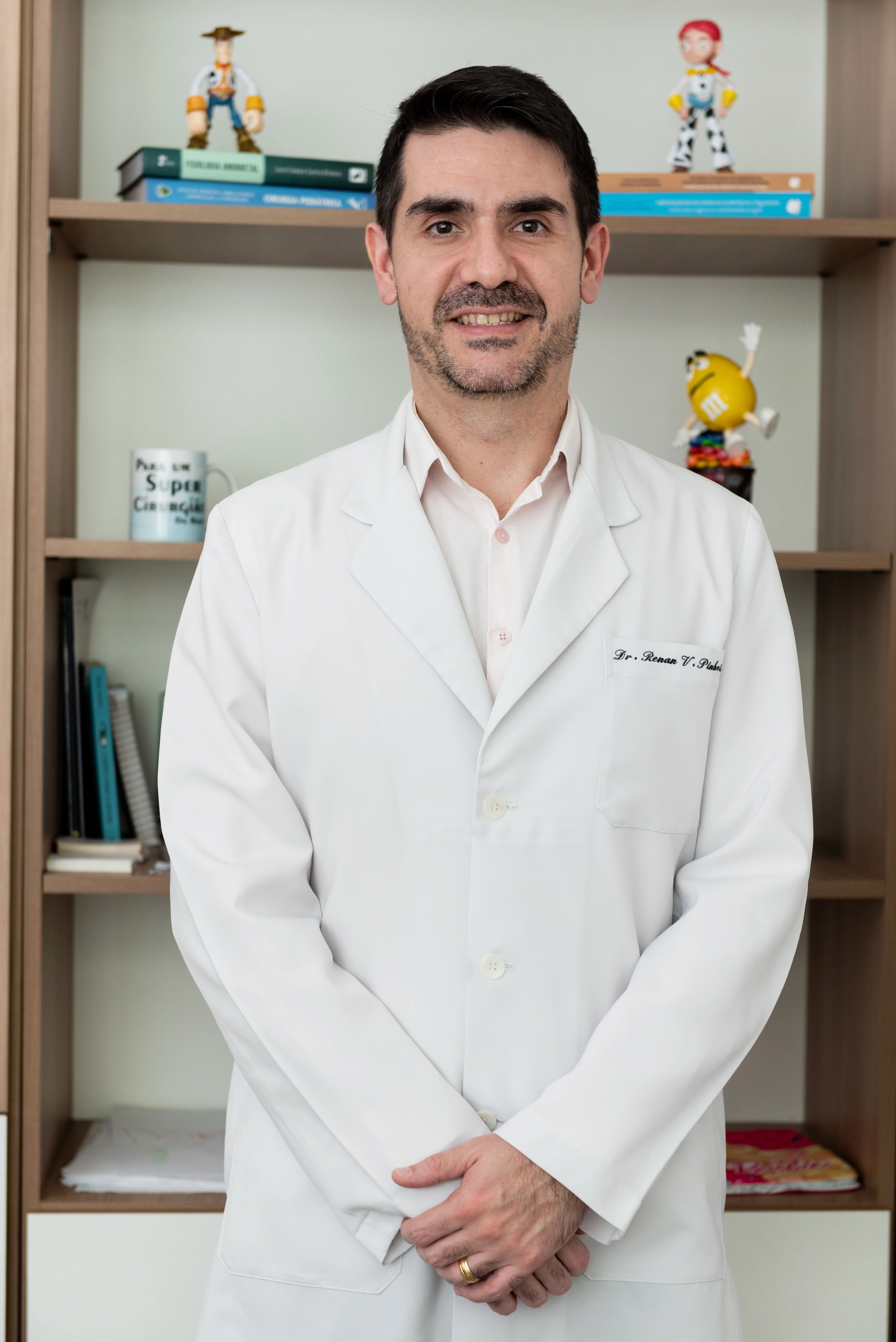 Foto profissional do Dr. Renan, um médico de barba rala, em pé, sorrindo e olhando para a câmera. Ele veste um jaleco branco sobre uma camisa social rosa. Atrás dele, em uma prateleira, há brinquedos infantis como Woody, Jessie e um M&M.