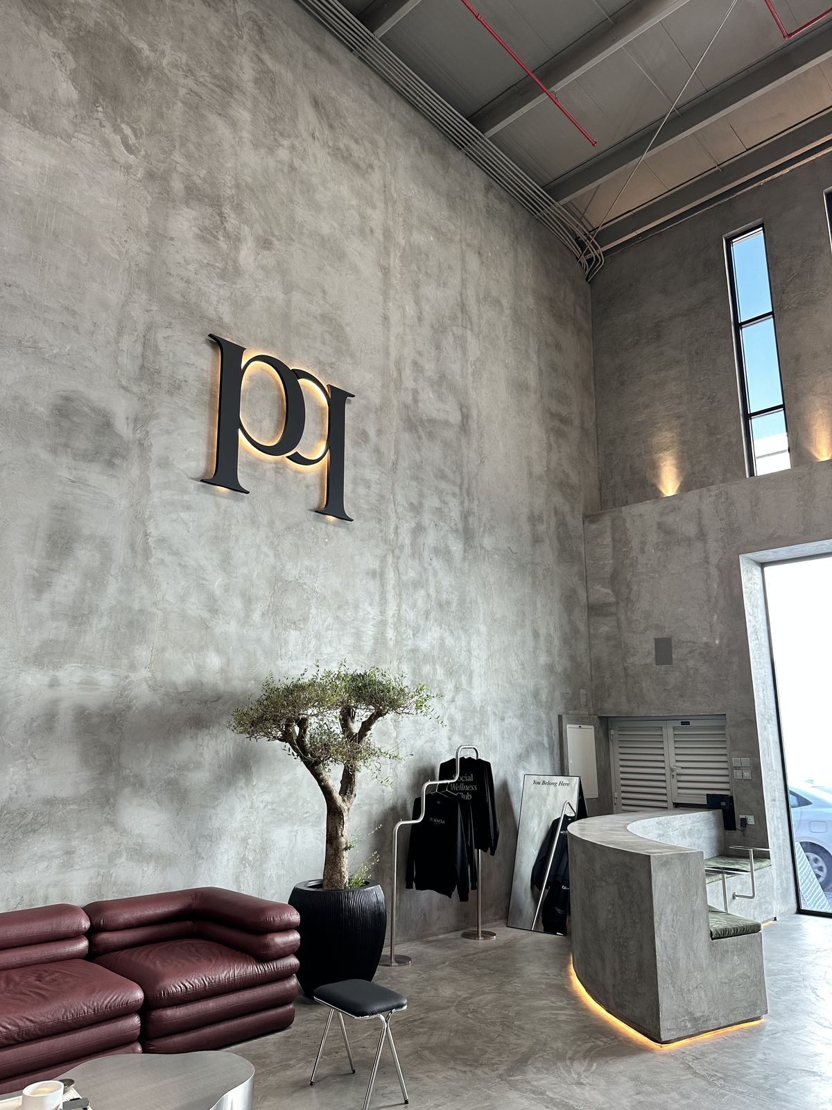 Modern Pilates studio reception with large illuminated “pl” dimensional wall letters mounted on a tall concrete wall above a minimalist reception desk and lounge seating.