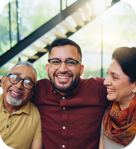 A warm, close-up portrait of a diverse family group: a young man smiling widely with his arm around two older adults