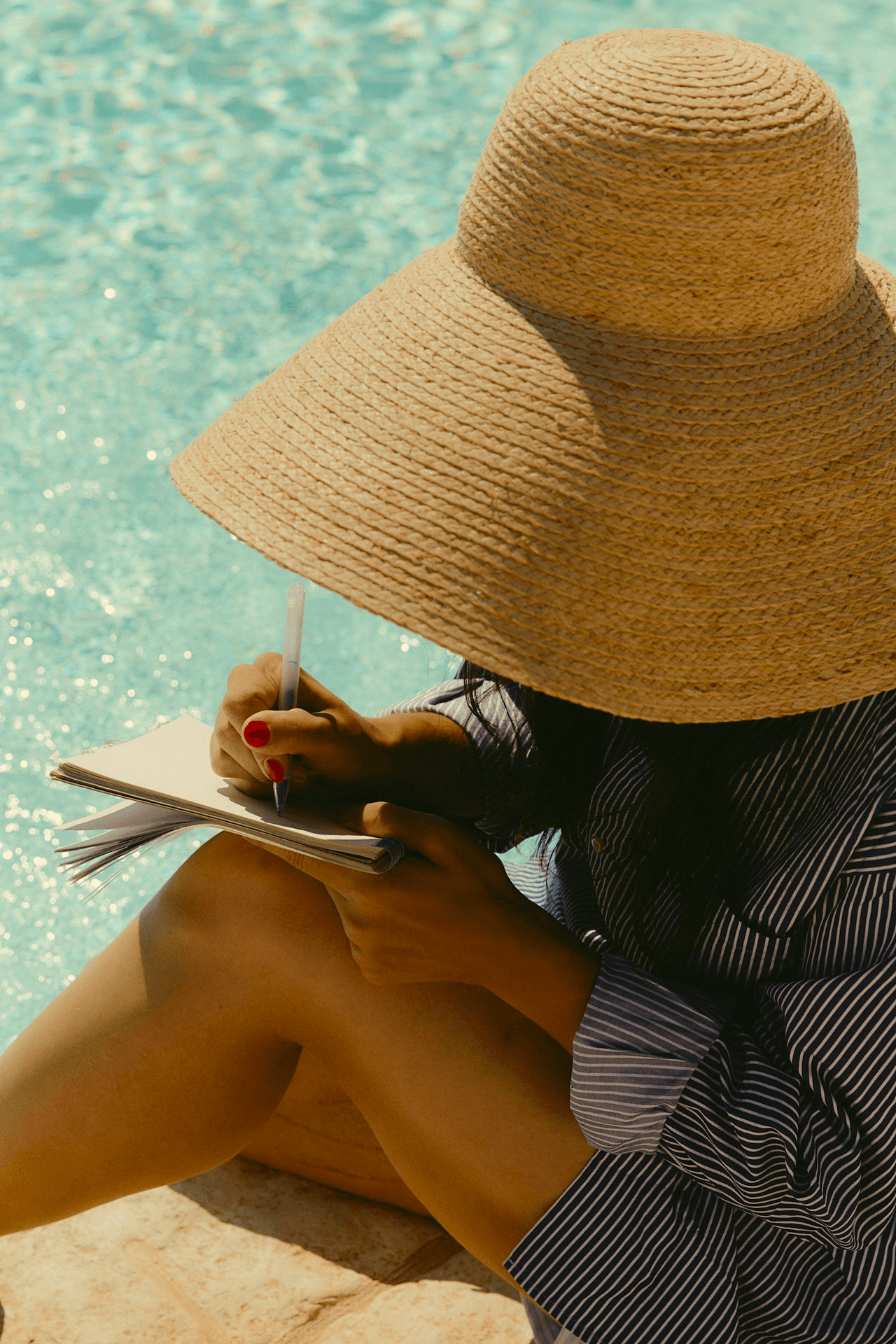 Une personne au grand chapeau de paille écrit dans un carnet près d'une piscine bleue. Ongles rouges.