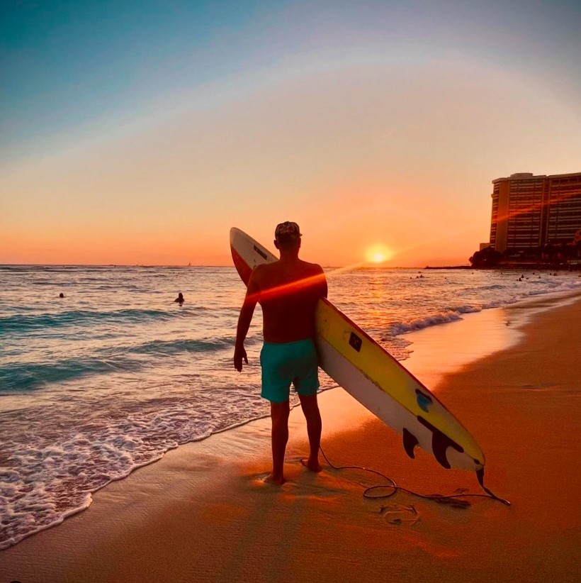 Kevin Welch stands barefoot at the shoreline in Waikiki holding a surfboard and looking toward a vivid ocean sunset after finishing a day of surfing with friends, with gentle waves and distant swimmers in the water and beachfront buildings glowing in the evening light.