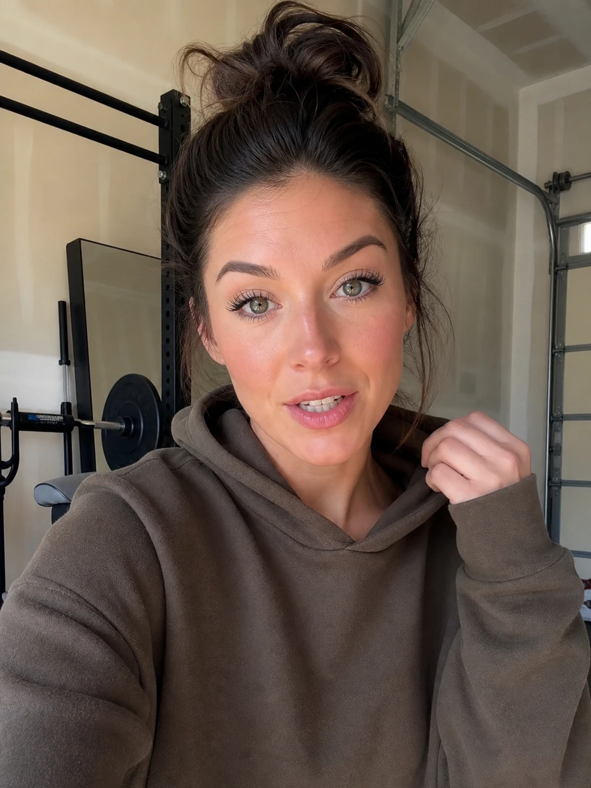 UGC-style photo: beauty creator, in a garage workout corner, trying on a plain hoodie while talking to camera, close vertical talking-head shot.