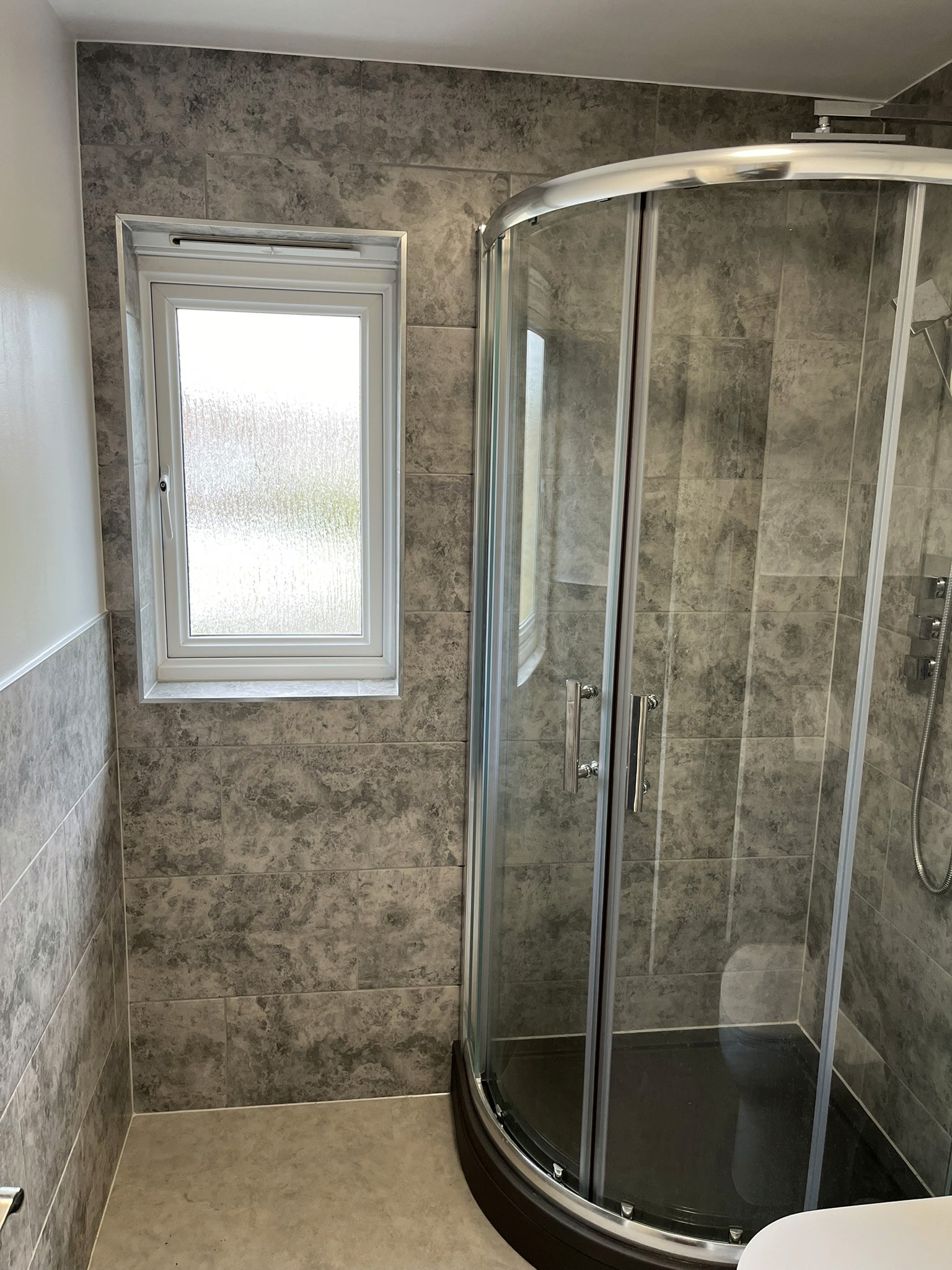 Finished shower enclosure with a glass door, grey stone-effect wall tiles and a frosted window.