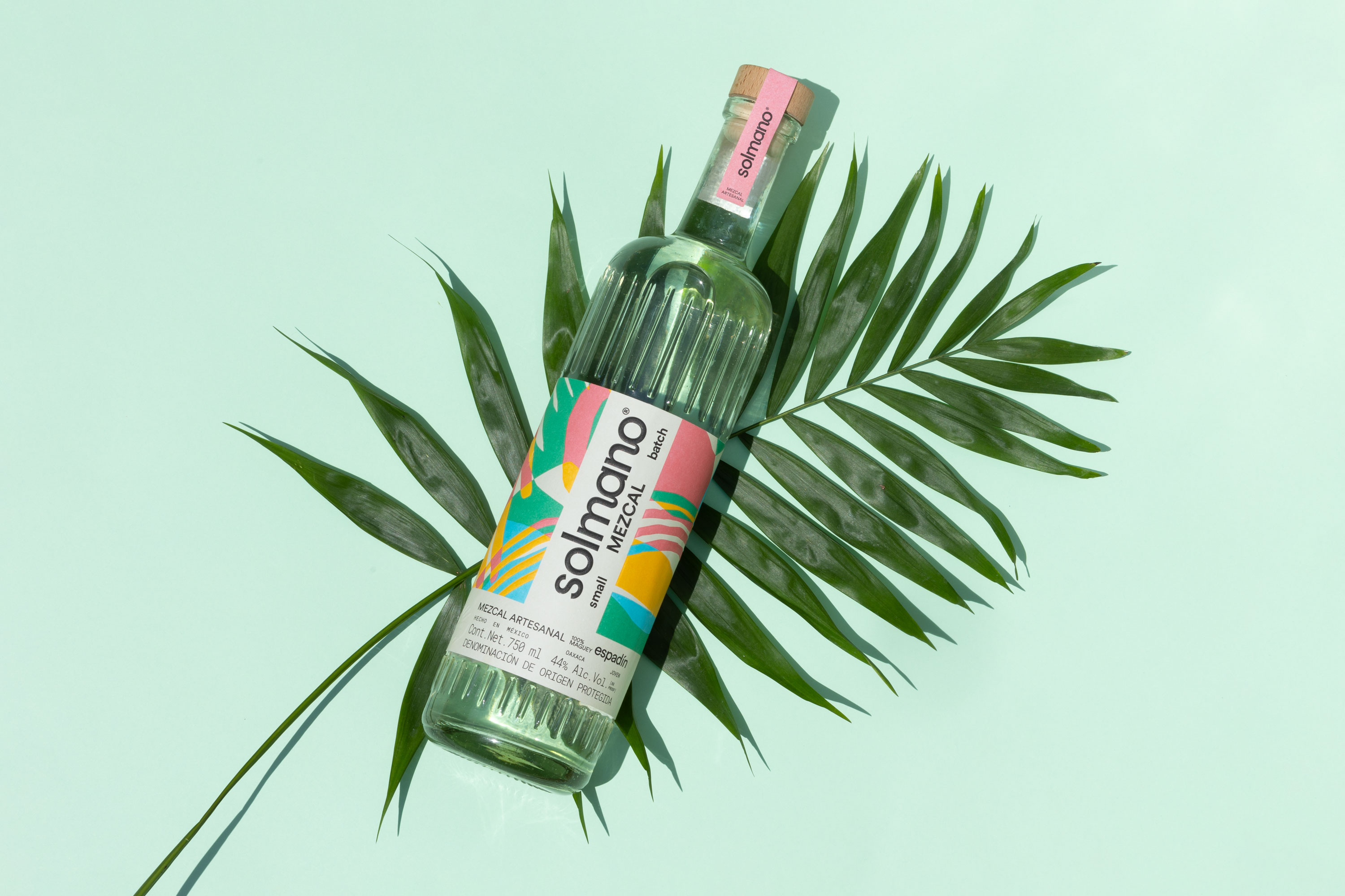 Solmano Espadin mezcal bottle resting on tropical palm leaves against a mint green background