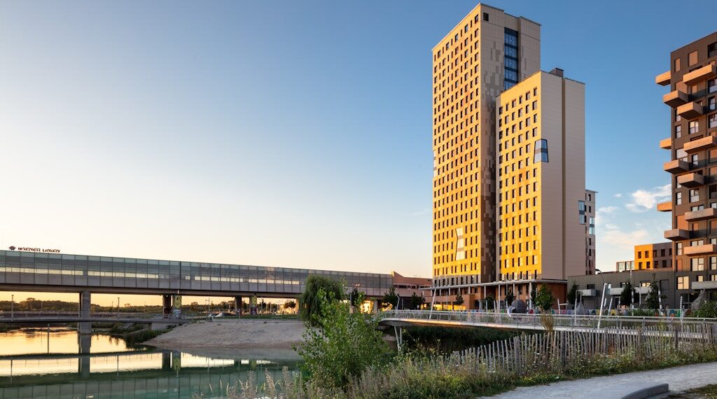 Modern red brick apartment or office building viewed from below against a blue sky, showcasing its geometric architecture with evenly spaced windows and balconies.