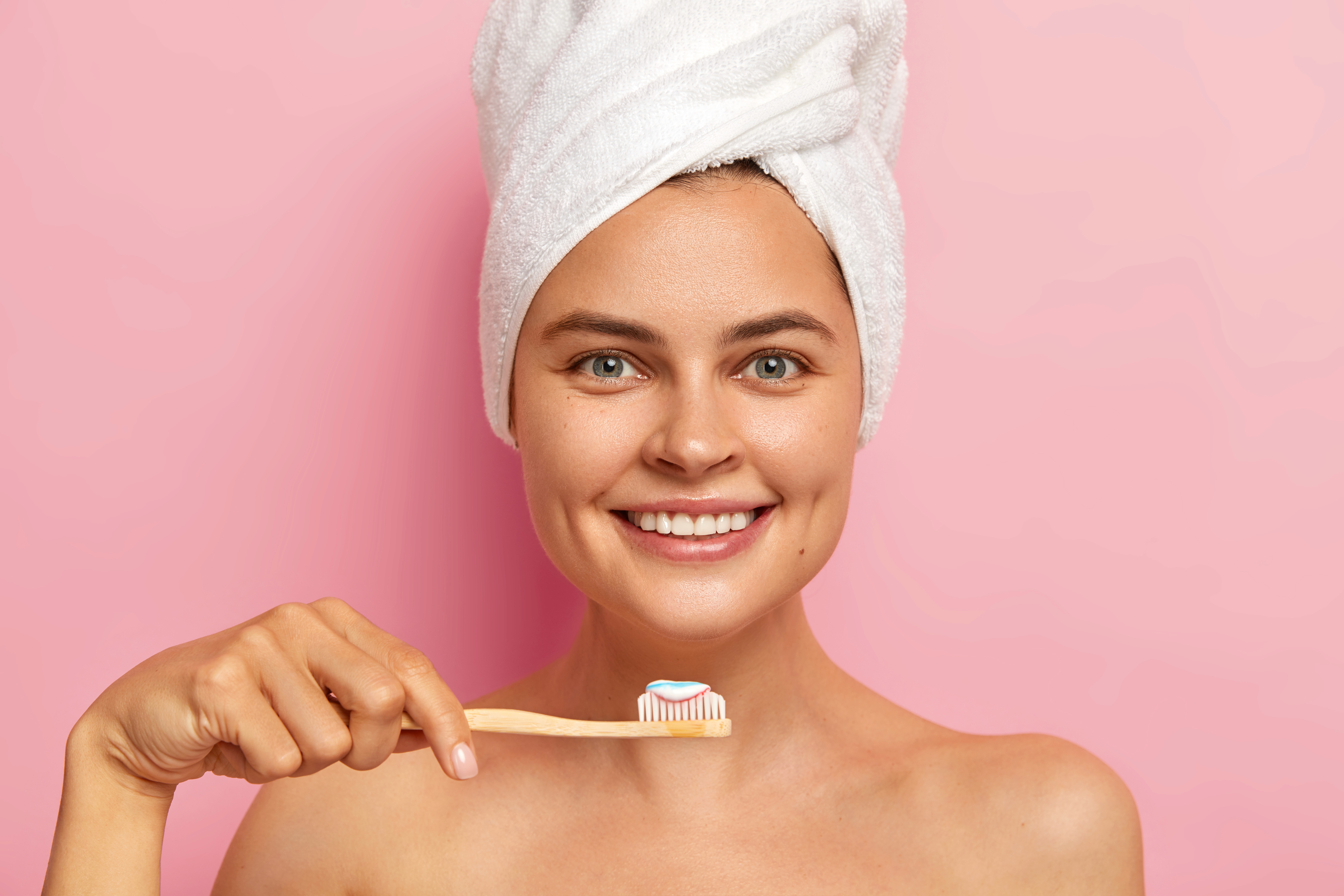 Smiling person with a towel on their head holding a bamboo toothbrush with toothpaste. They stand against a pink background. The tone is fresh and cheerful.